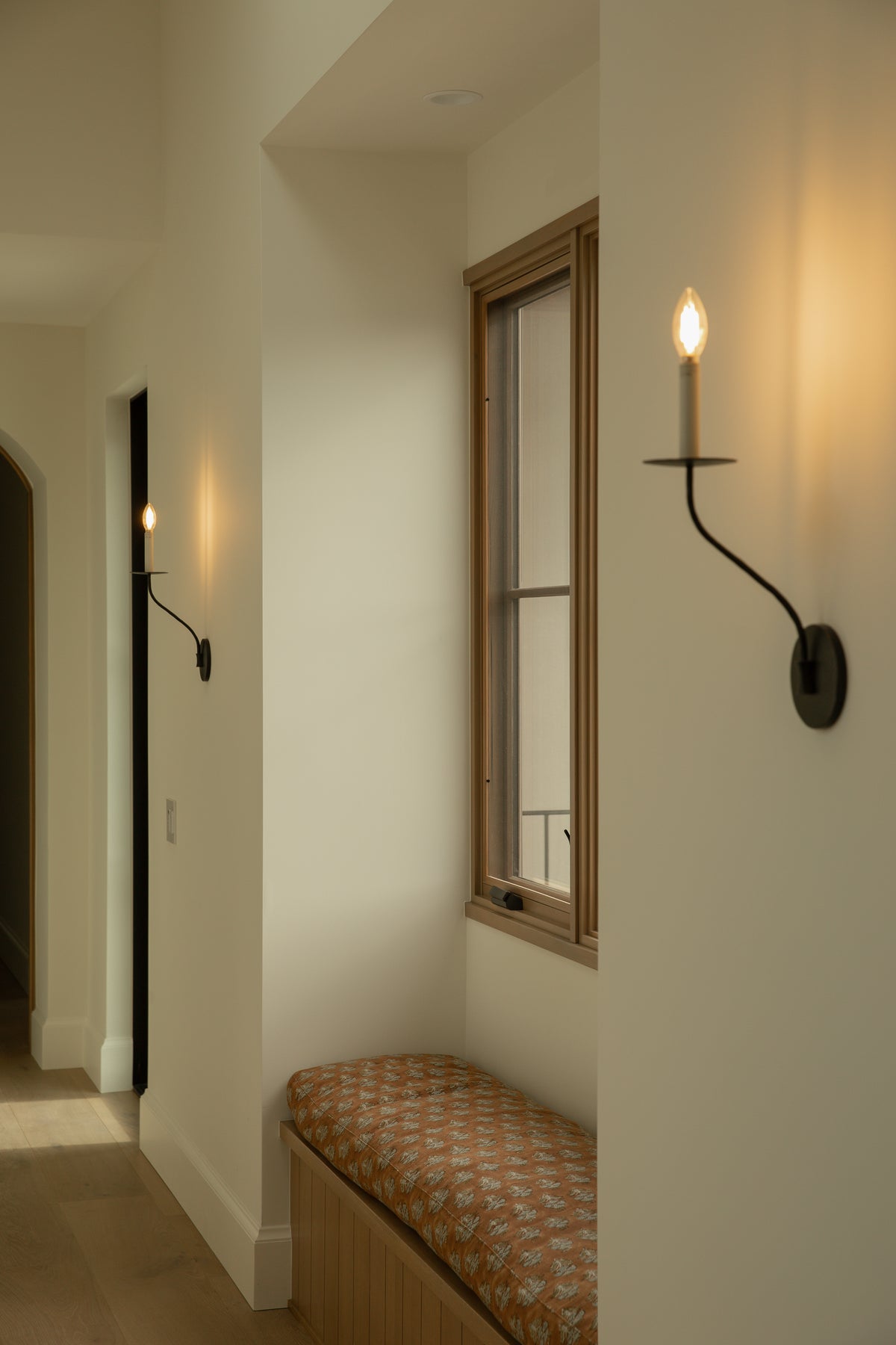 A bright hallway nook featuring a built-in window seat with a patterned cushion, framed by classic black iron candle wall sconces, crisp white walls, and a wood-framed window.