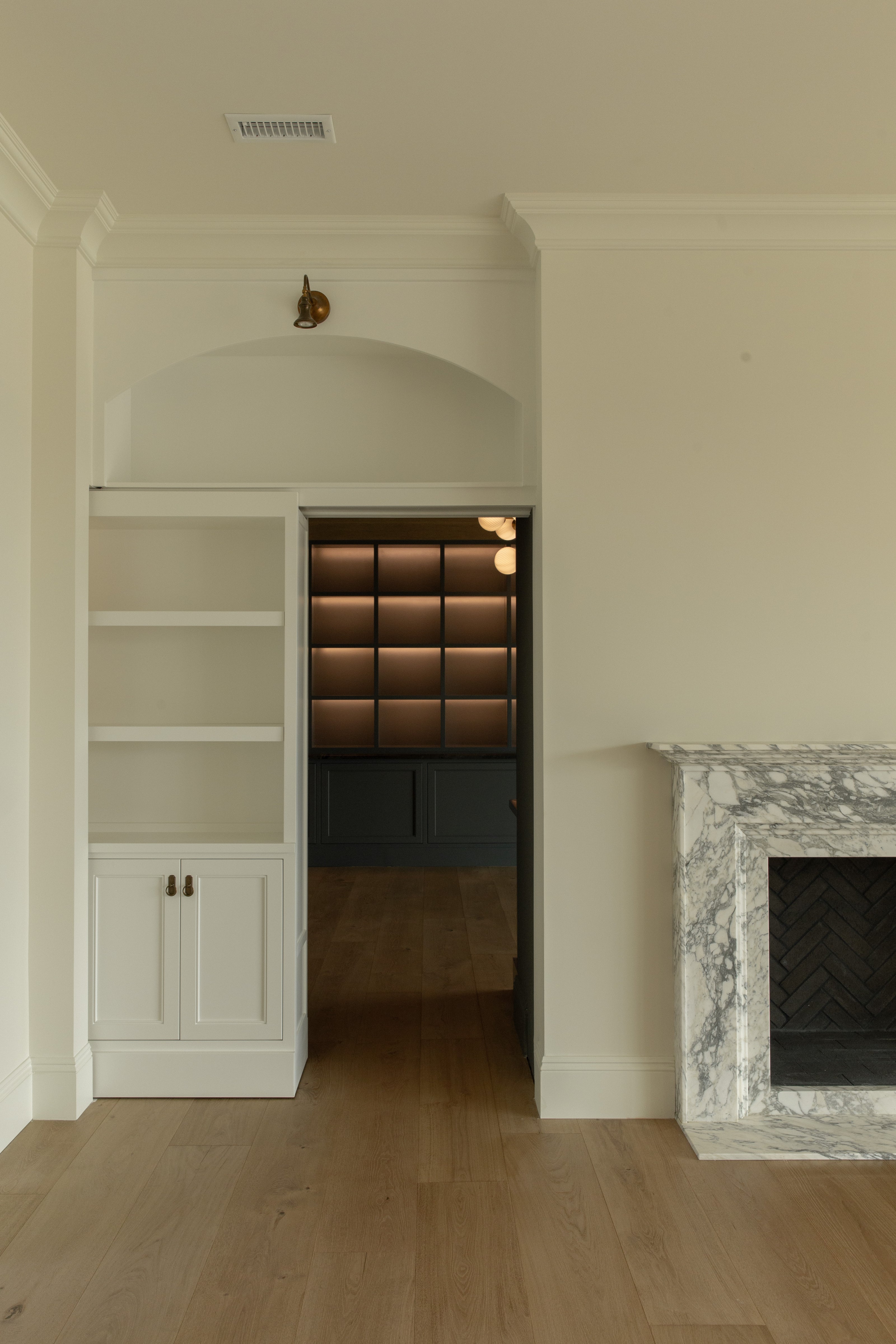A white built-in bookcase functioning as a hidden sliding door, revealing a dark and moody speakeasy bar inside a Rossmoor living room next to a marble fireplace.