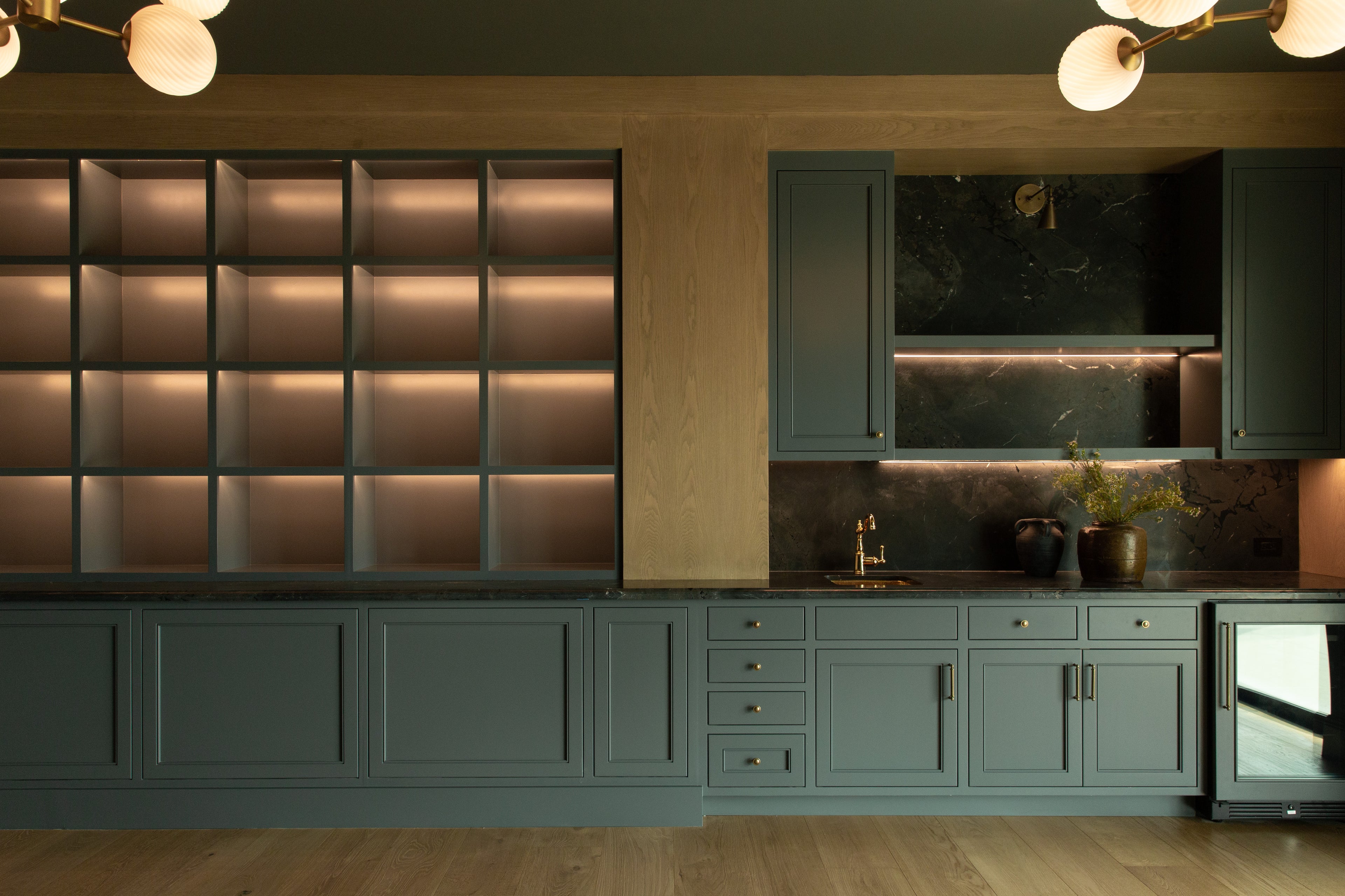 Straight-on view of a custom home bar in Rossmoor, showcasing extensive lit display shelving, dark green lower cabinets, and a bar sink with a brass faucet against a dark marble backsplash.