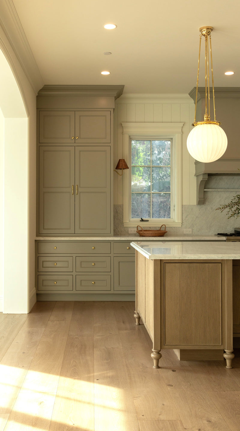 Detail of a custom wood kitchen island with ribbed paneling and classic turned legs, positioned in front of floor-to-ceiling olive green pigeon pantry cabinets and a window with a brass wall sconce.