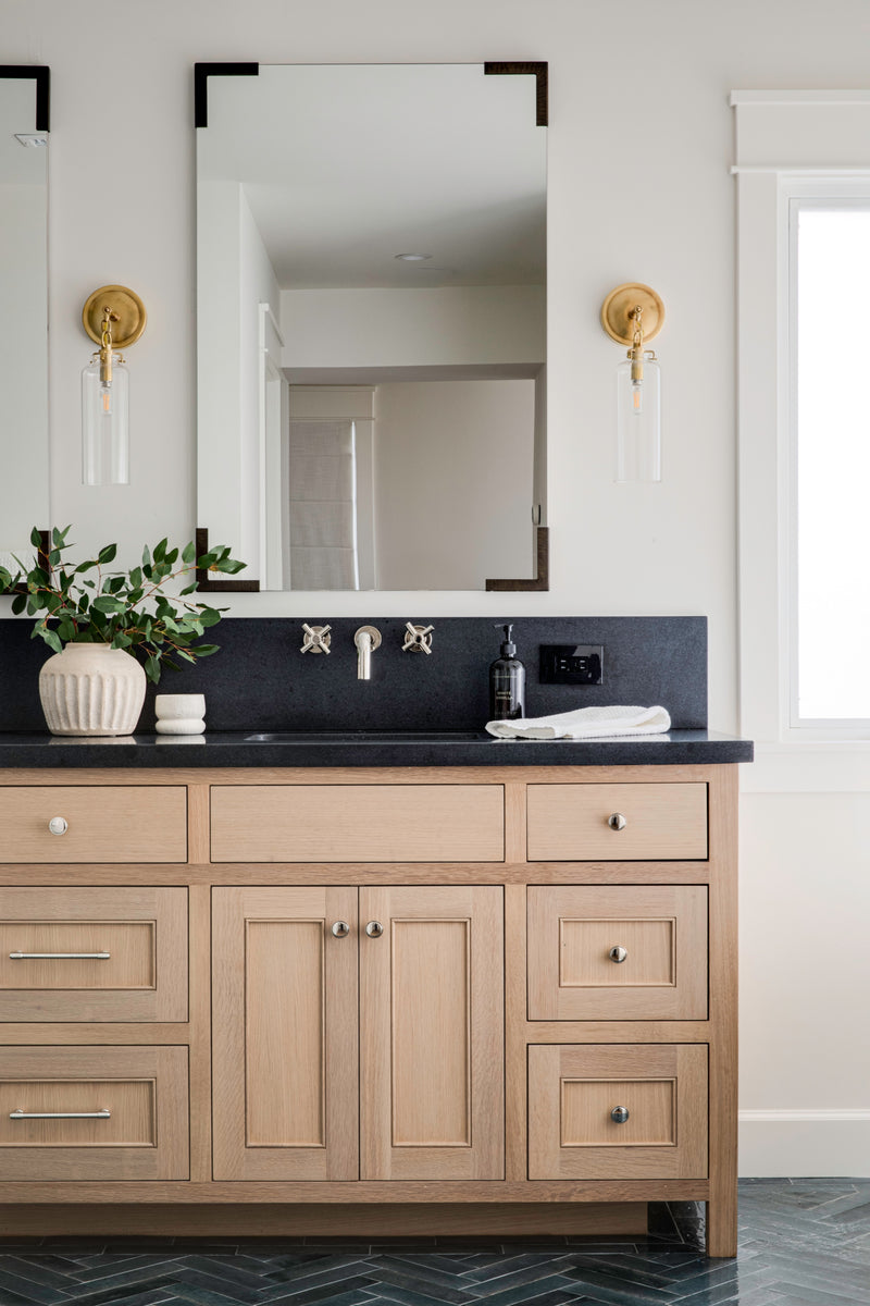 Light oak double vanity featuring a contrasting dark stone countertop, brass wall sconces, and refined cabinetry hardware in a custom bathroom design.