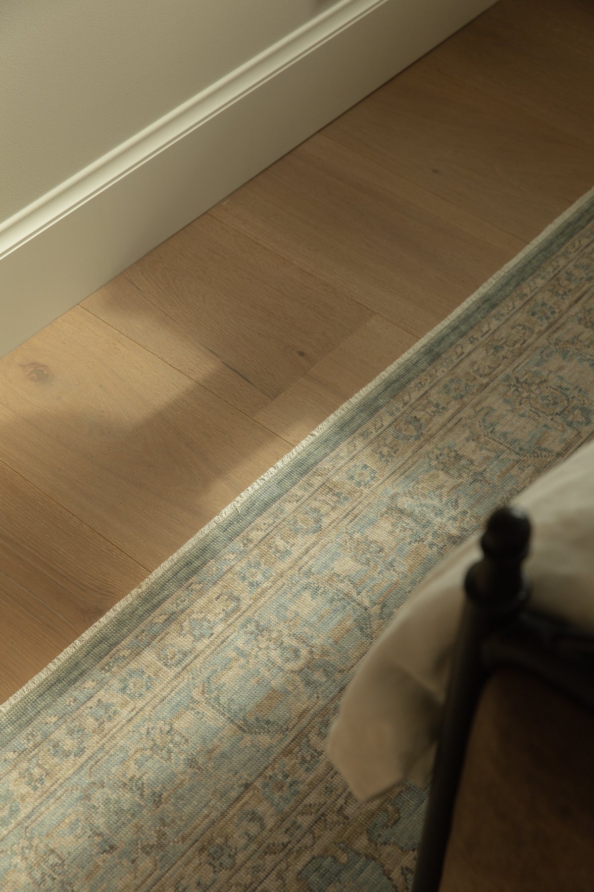 Architectural detail shot showing wide-plank light oak hardwood flooring meeting crisp white baseboards, beautifully accented by a vintage-inspired blue and neutral patterned area rug near an iron bed frame.