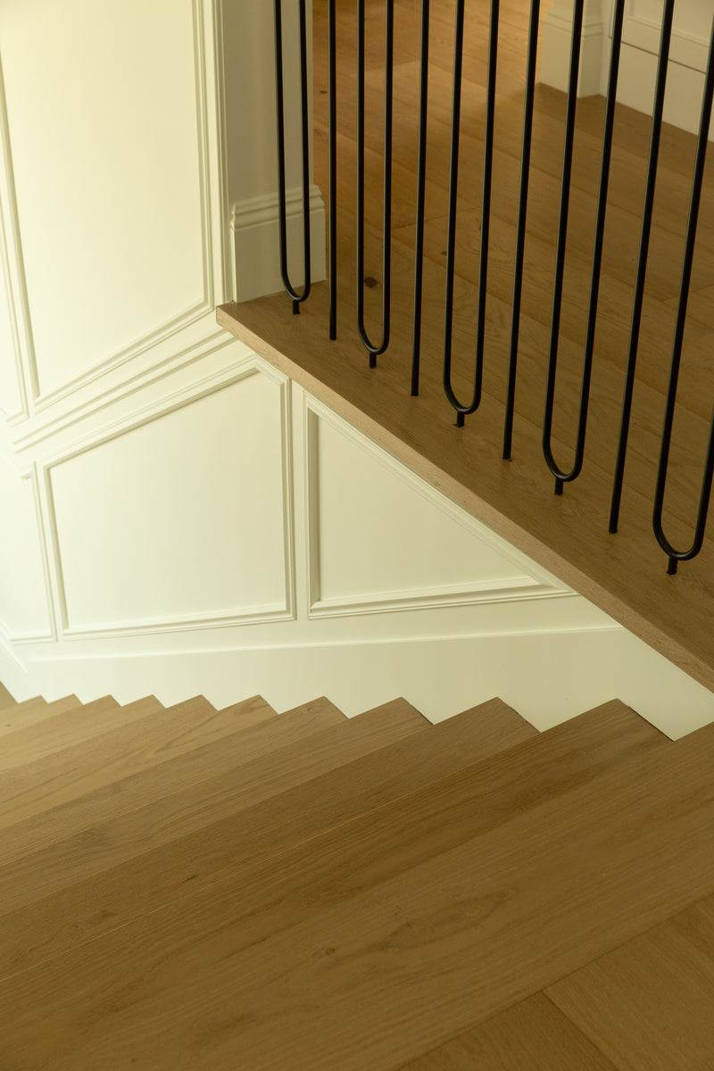 View looking down a light oak wood staircase featuring modern black iron balusters and classic white wainscoting wall details in a Rossmoor Tudor home rebuild.