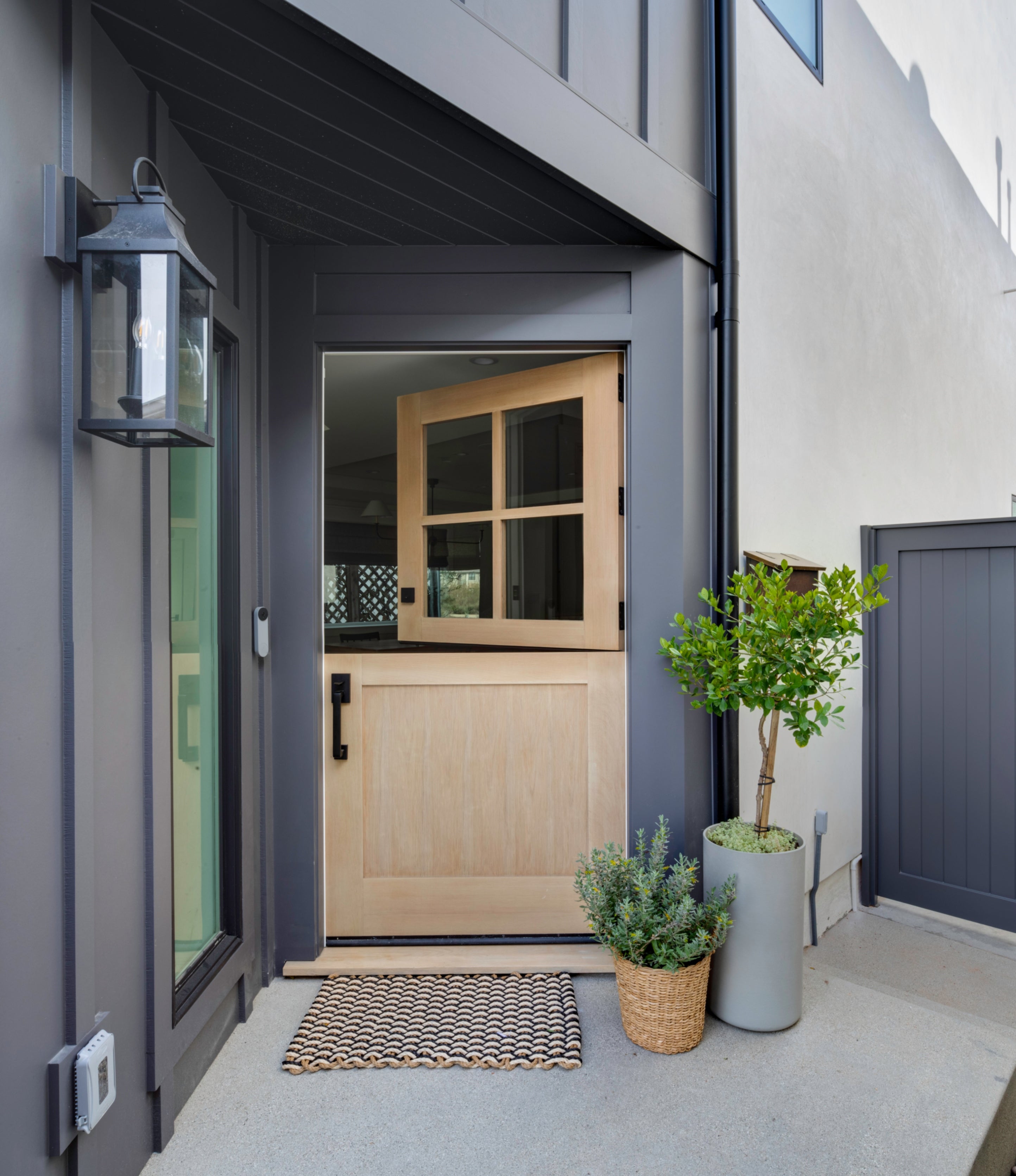 Custom light wood Dutch door entry by B Farm Design, featuring modern black hardware, a classic lantern-style sconce, and minimalist exterior finishes.