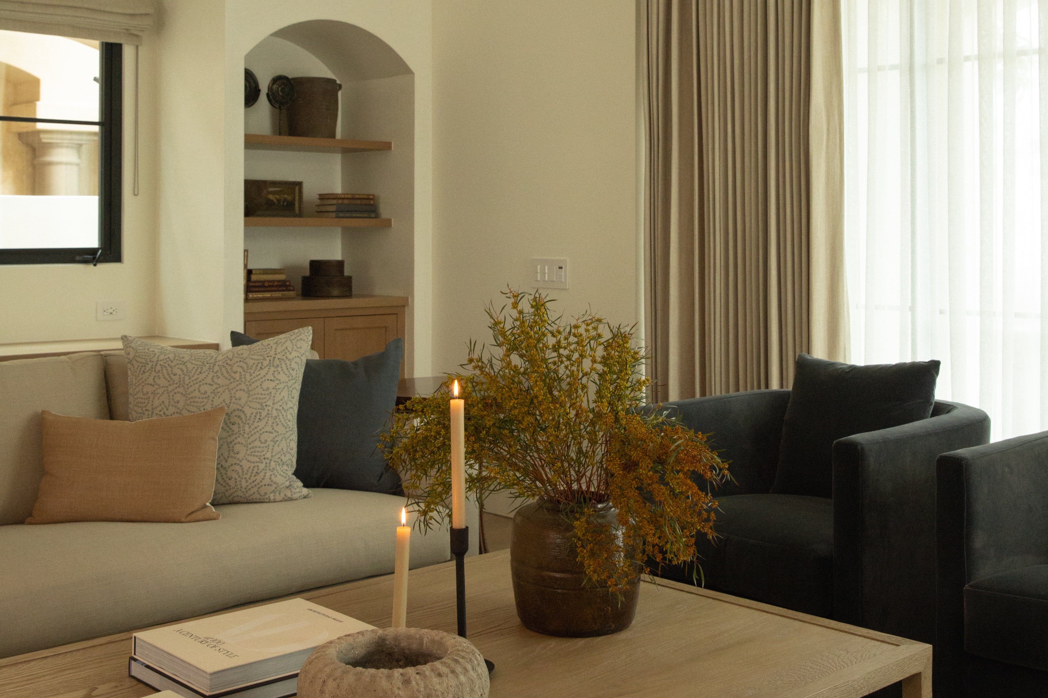 A transitional living room featuring a custom arched built-in wall niche with wood shelving, a light beige sofa, dark velvet armchairs, and floor-to-ceiling pinch-pleat linen drapery.