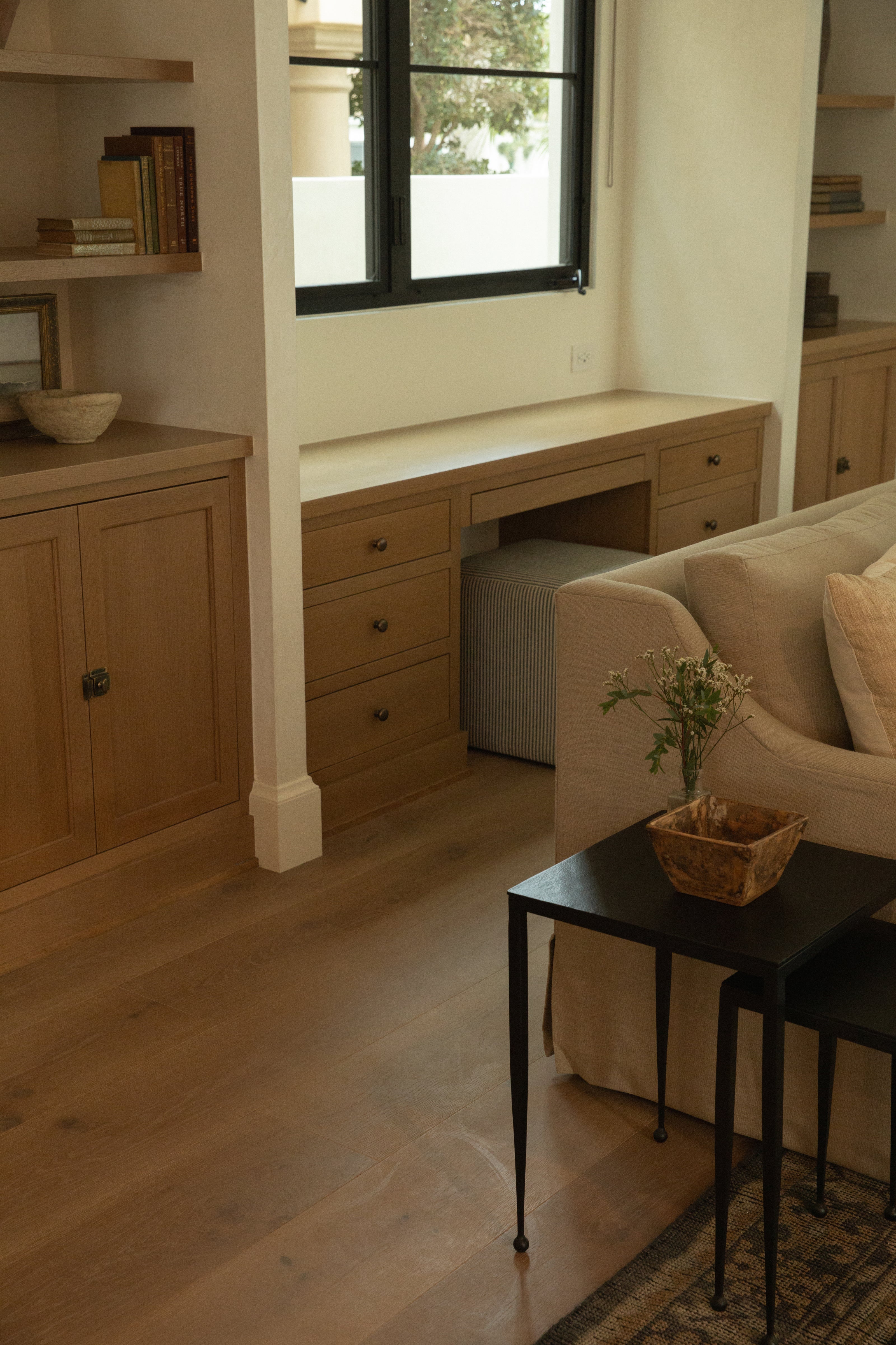Custom light oak built-in desk and drawer cabinetry tucked perfectly into a living room window alcove, paired with wide-plank hardwood floors and a comfortable linen sofa.