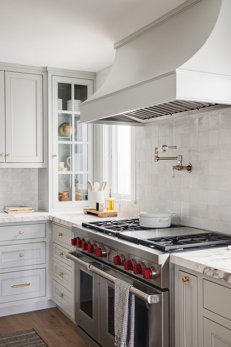 Luxury kitchen remodel featuring a custom-built range hood, professional-grade stainless steel range with signature red knobs, and elegant marble countertops.