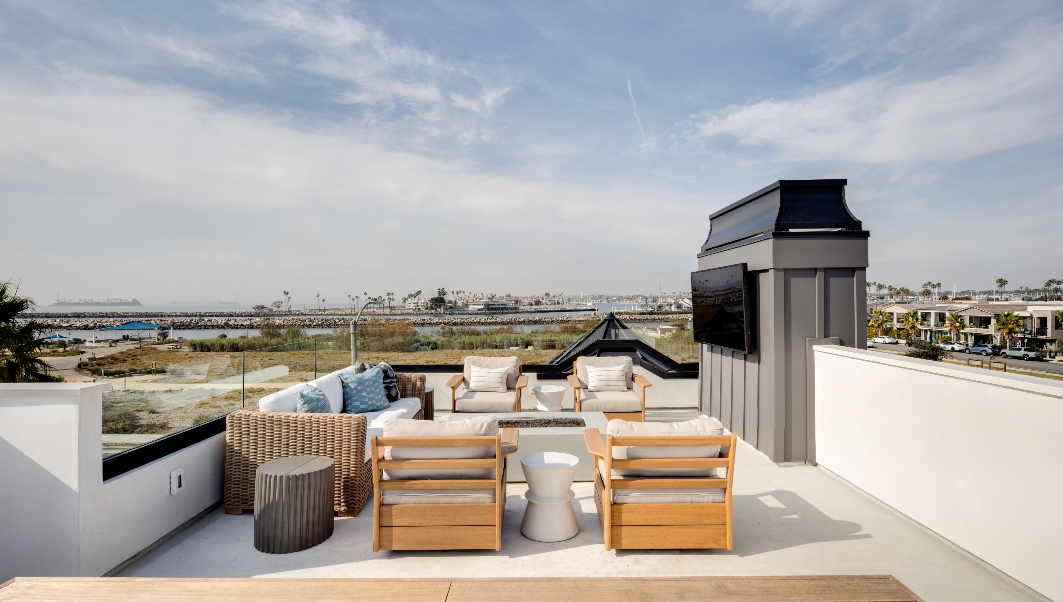 Cozy and modern rooftop lounge with an extensive view. Weathered wicker sofa and four low-profile wooden club chairs are arranged around a square fire pit. Features blue patterned pillows, unique cylindrical end tables, and an outdoor television mounted on a textured charcoal gray fireplace chimney structure.