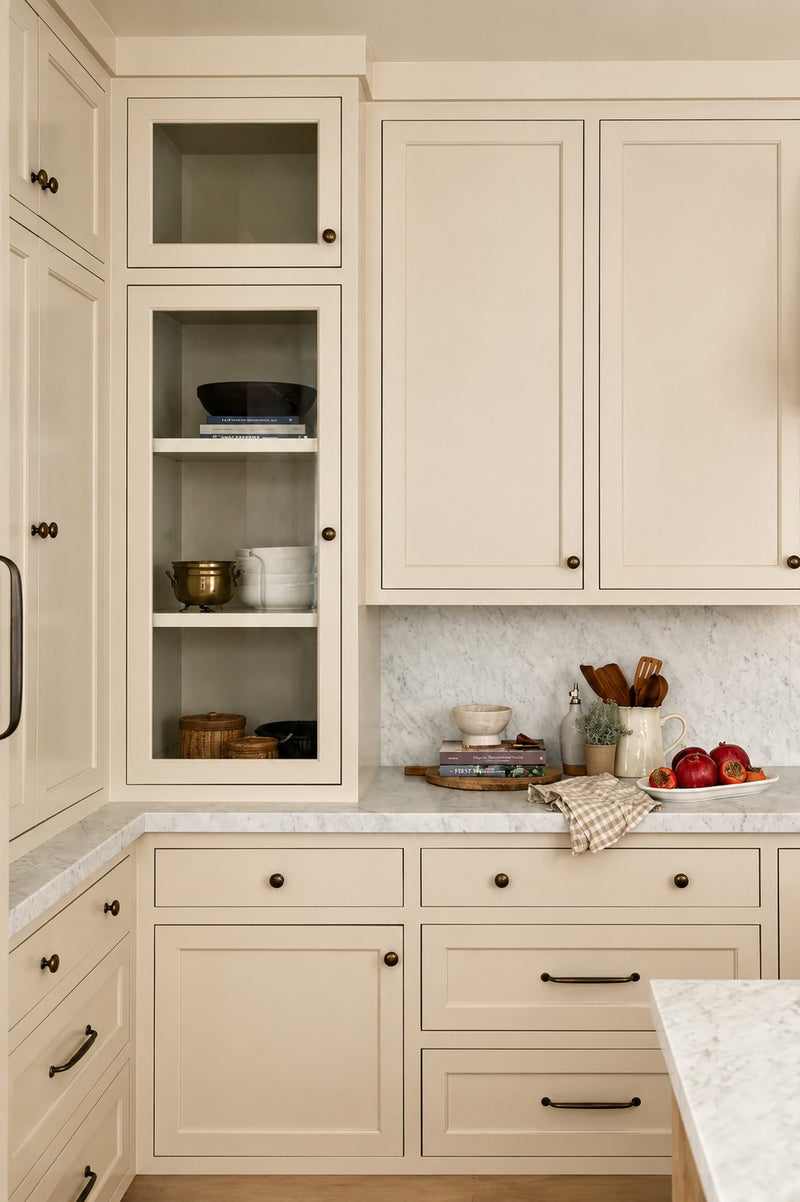 A beautifully styled coastal farmhouse kitchen corner featuring cream shaker cabinetry, a tall glass-front display cabinet, and a solid marble slab backsplash decorated with fresh fruit and wooden cooking utensils.