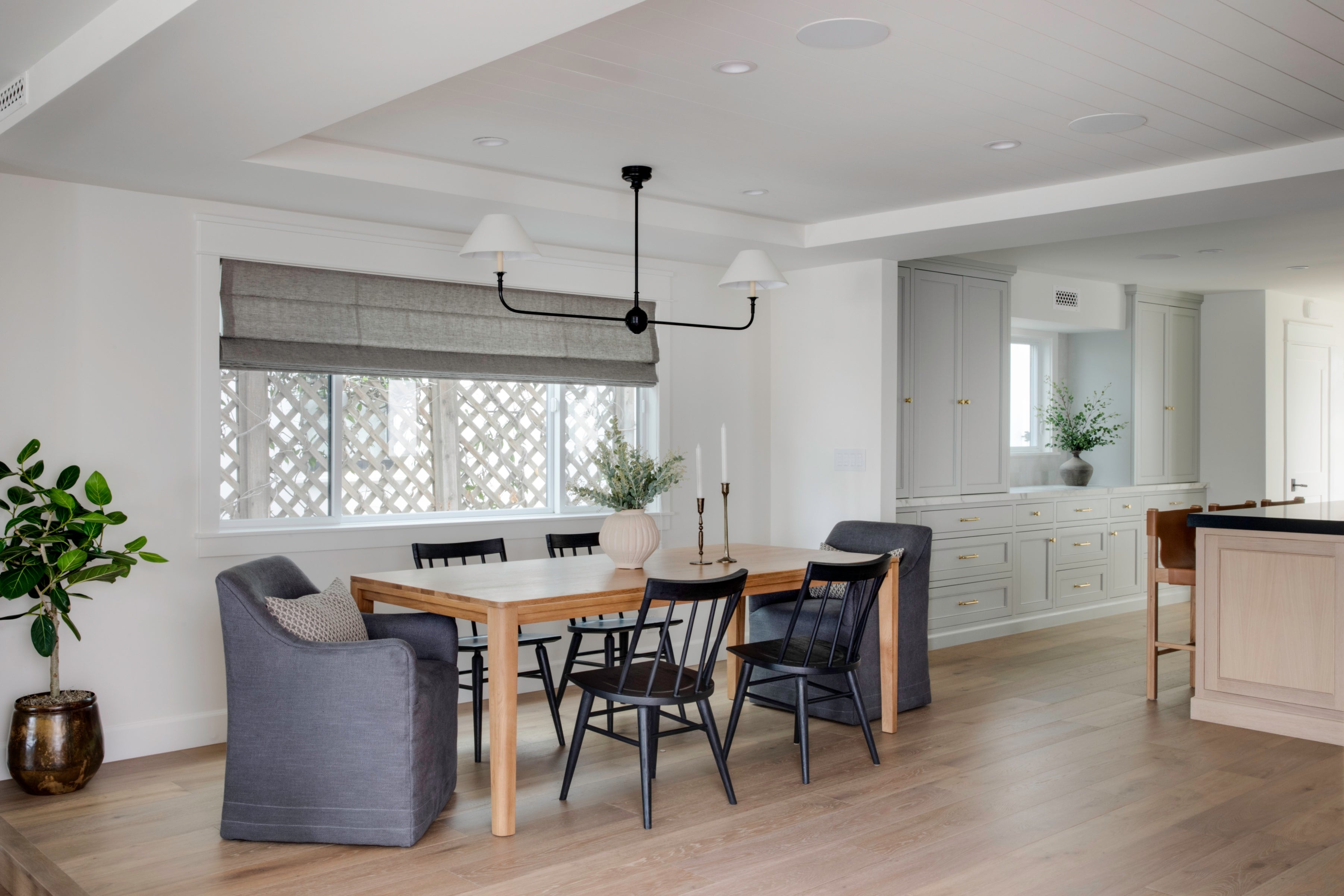 An open-concept modern coastal dining room designed by a B Farm Design in Seal Beach. The space features a light wood dining table with black spindle chairs and gray slipcovered armchairs, sitting beneath a black linear chandelier with white shades. In the background, custom light pigeon kitchen cabinetry with brass hardware is visible over light oak flooring