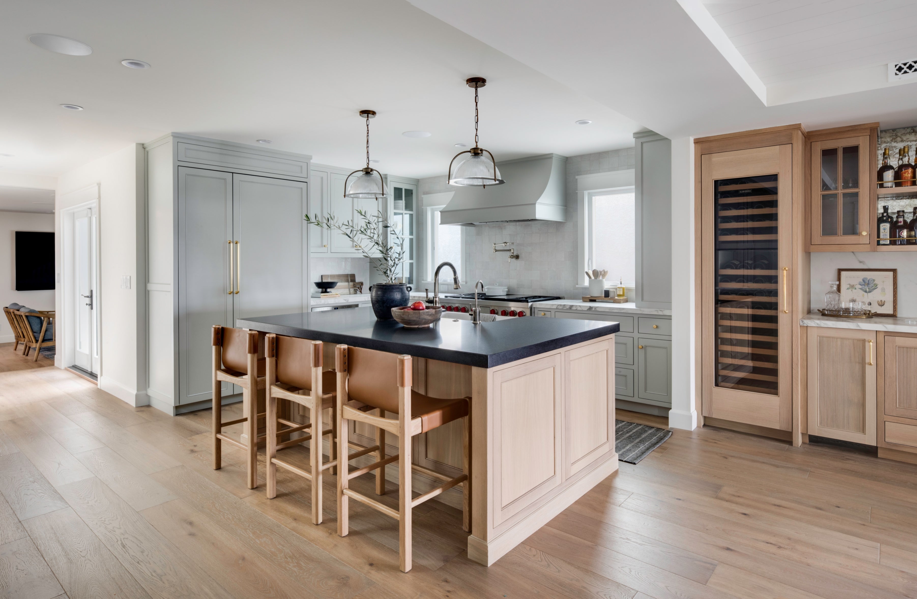 Modern coastal kitchen in an Old Town Seal Beach custom build, featuring bespoke cabinetry in sage green and natural white oak, a central island with a contrasting dark stone countertop, leather-upholstered bar stools, and a built-in wine refrigerator.
