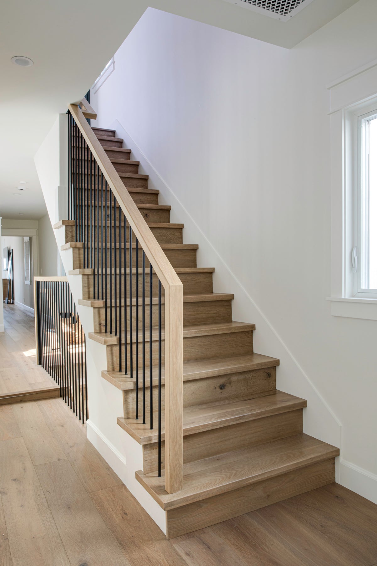 Modern staircase remodel featuring warm oak wood treads, a simple handrail, and sleek vertical black metal balusters in a white-walled home interior.