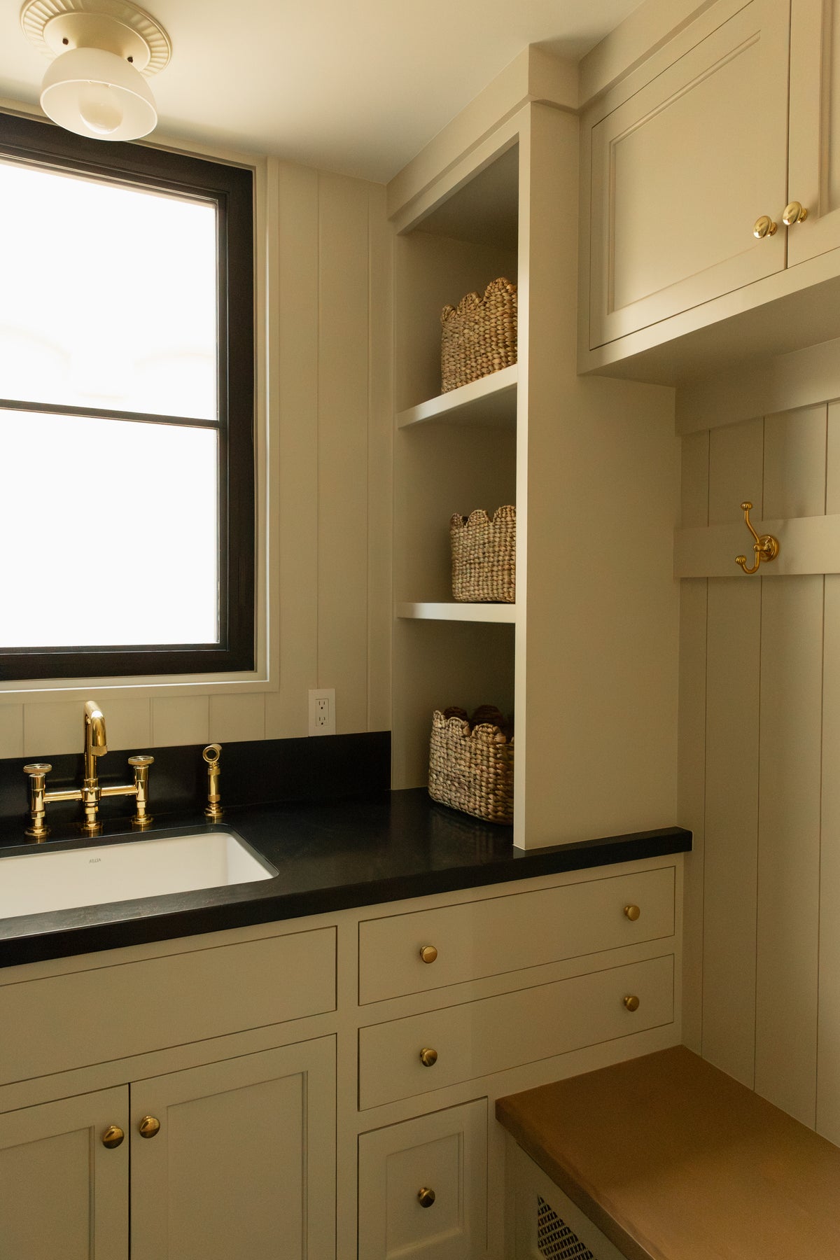 Close-up view of custom transitional mudroom storage featuring unlacquered brass sconces, a dark counter over an off-white cabinet, and neatly styled woven baskets.