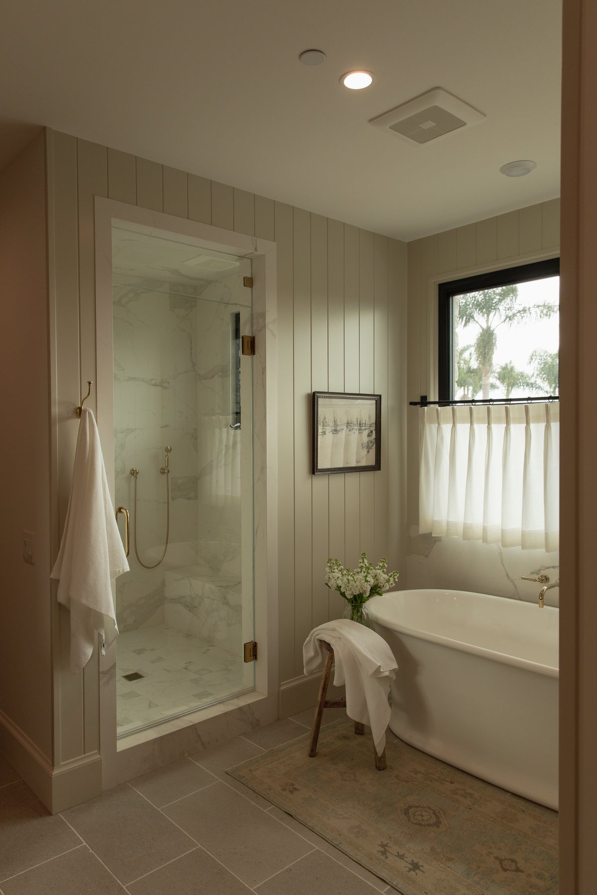 A stunning transitional villa bathroom in Old Town Seal Beach featuring a seamless glass walk-in shower with large marble tile, vertical painted wall paneling, and a classic white freestanding soaking tub.
