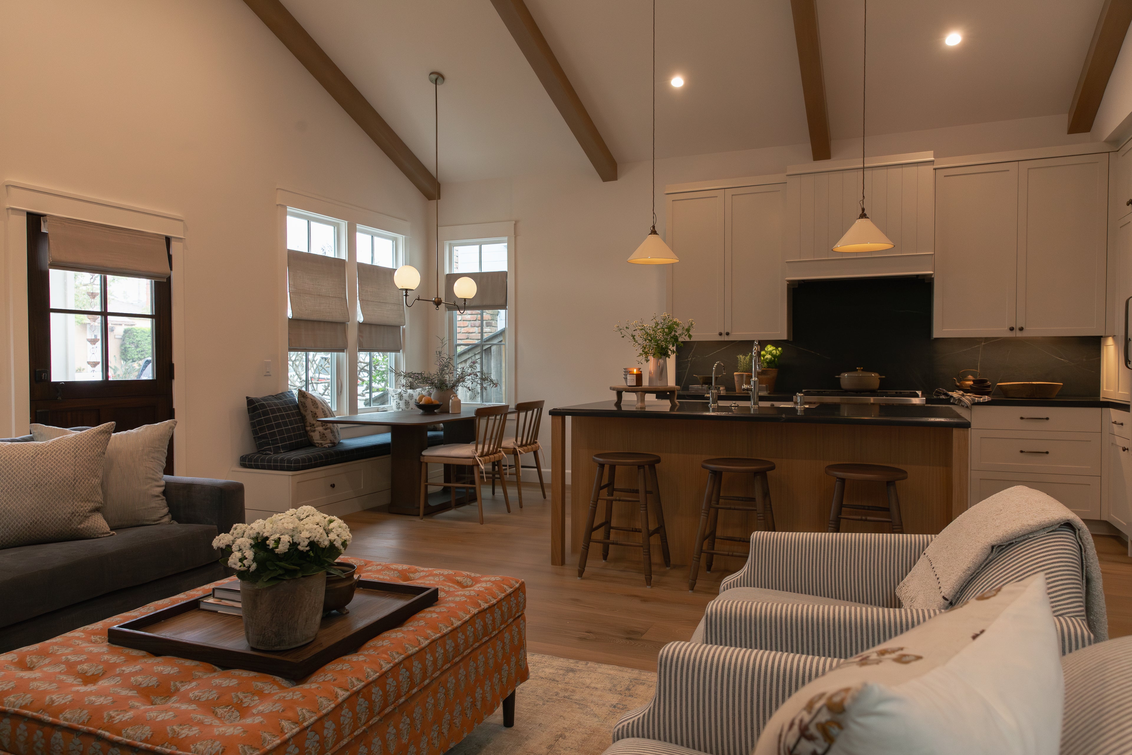 Wide view of an open-concept Seal Beach bungalow remodel demonstrating cohesive interior design, flowing from a transitional living room with striped chairs and an orange patterned ottoman directly into a custom kitchen with a built-in window seating area.