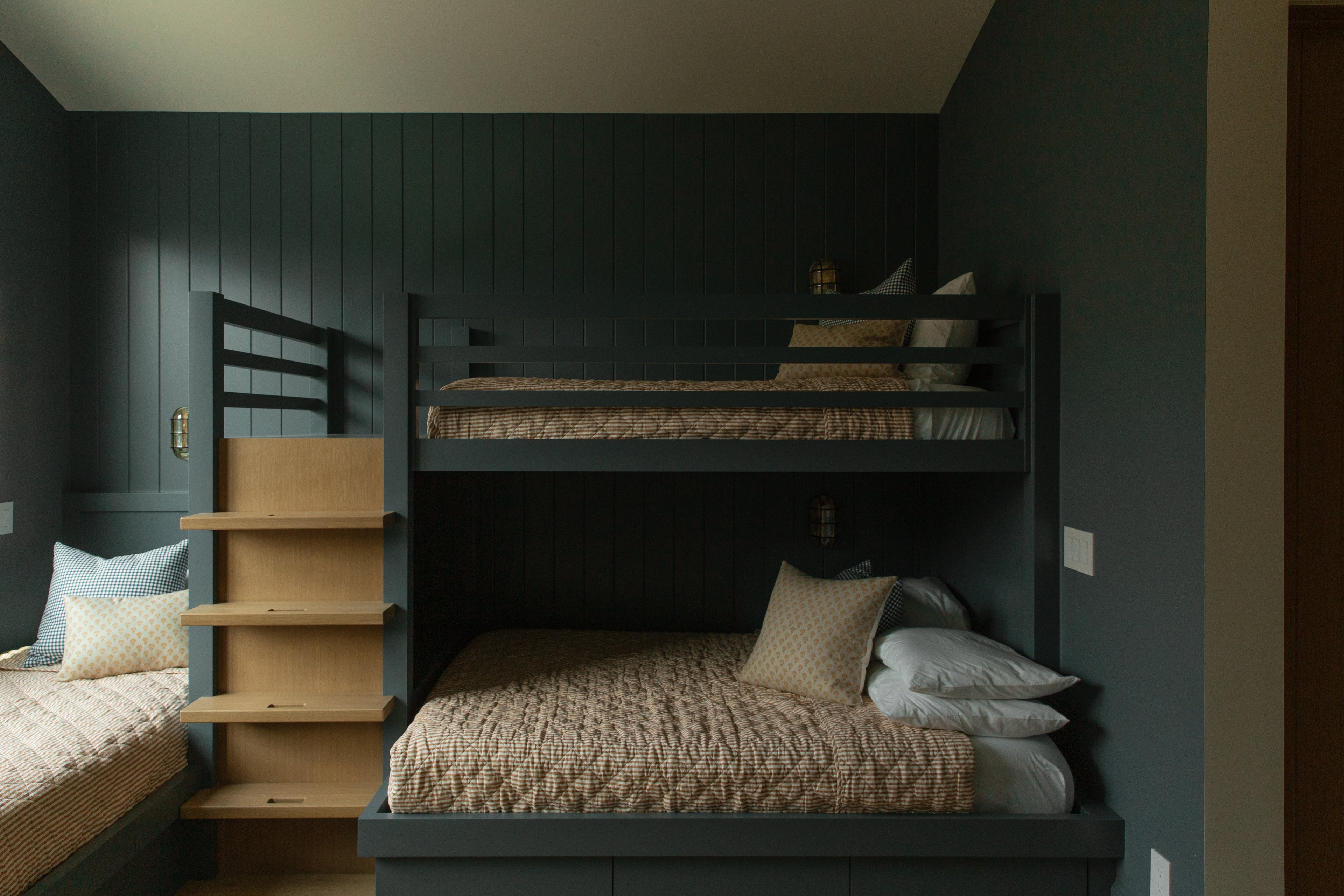 A custom-built kids bunk room in an Orange County transitional villa, featuring moody blue-grey vertical paneled walls, built-in bunk beds, and warm oak storage stairs.