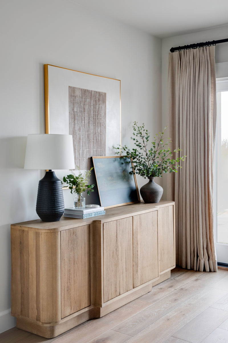 Interior design detail showcasing a natural wood console table styled in an organic modern aesthetic with neutral curtains and layered decorative accessories.