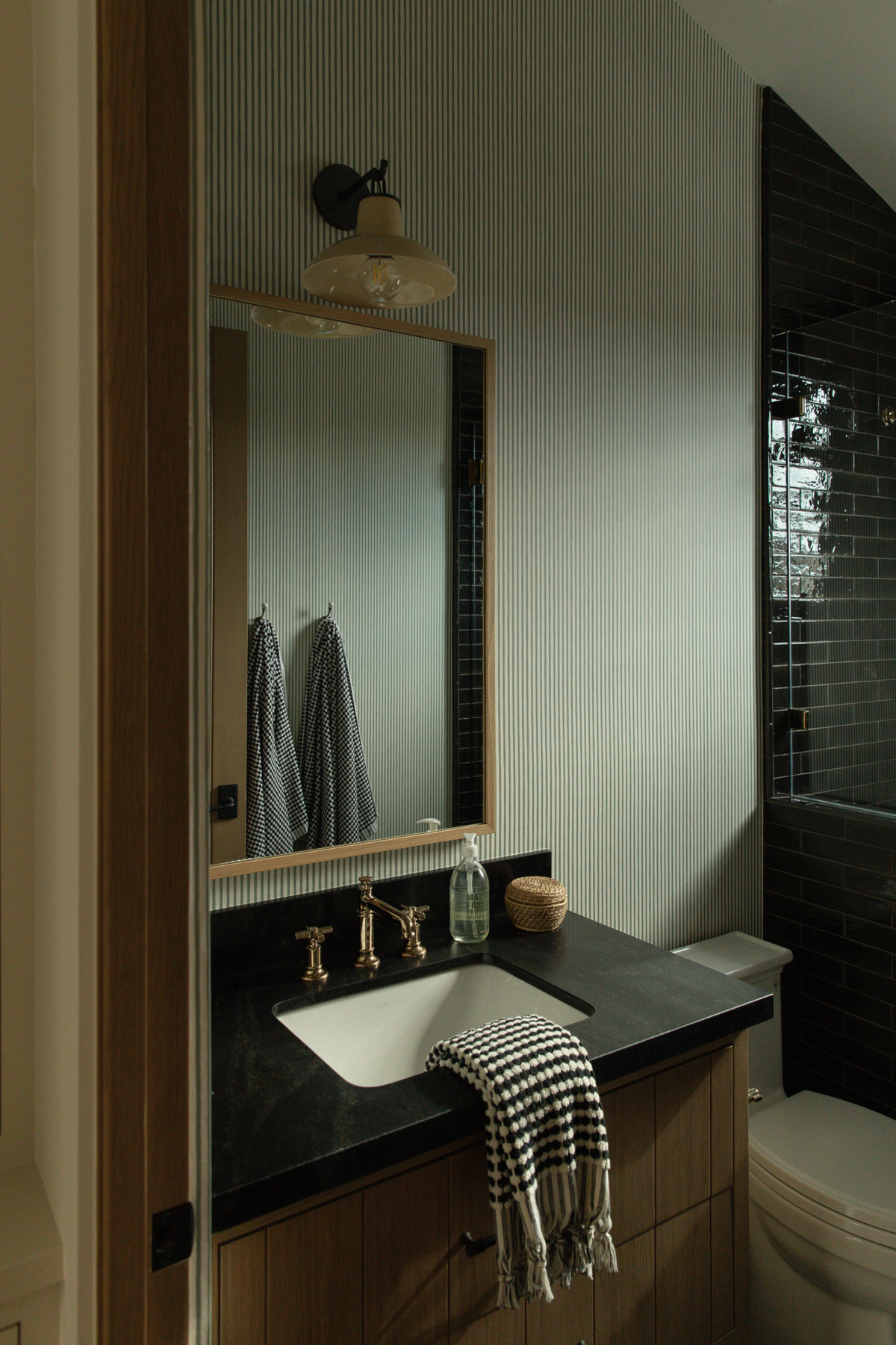 A transitional bathroom vanity featuring subtle pinstripe wallpaper, a dark stone countertop with a white undermount sink, unlacquered brass cross-handle faucets, and a custom wood cabinet.