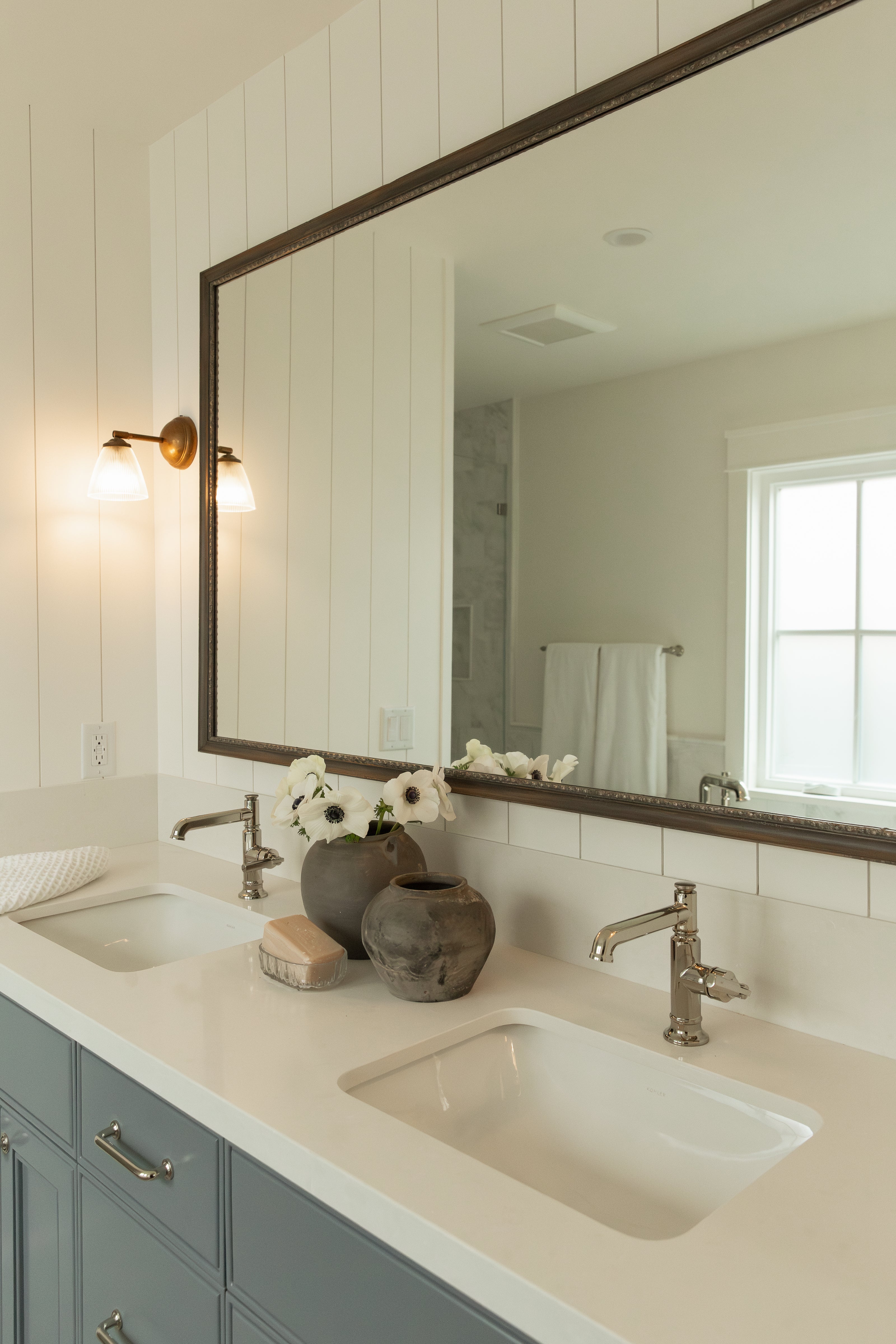 A primary bathroom redesign in Seal Beach featuring a custom slate-blue double vanity, white solid surface countertops, polished nickel faucets, and a large wood-framed mirror flanked by vintage-style sconces.