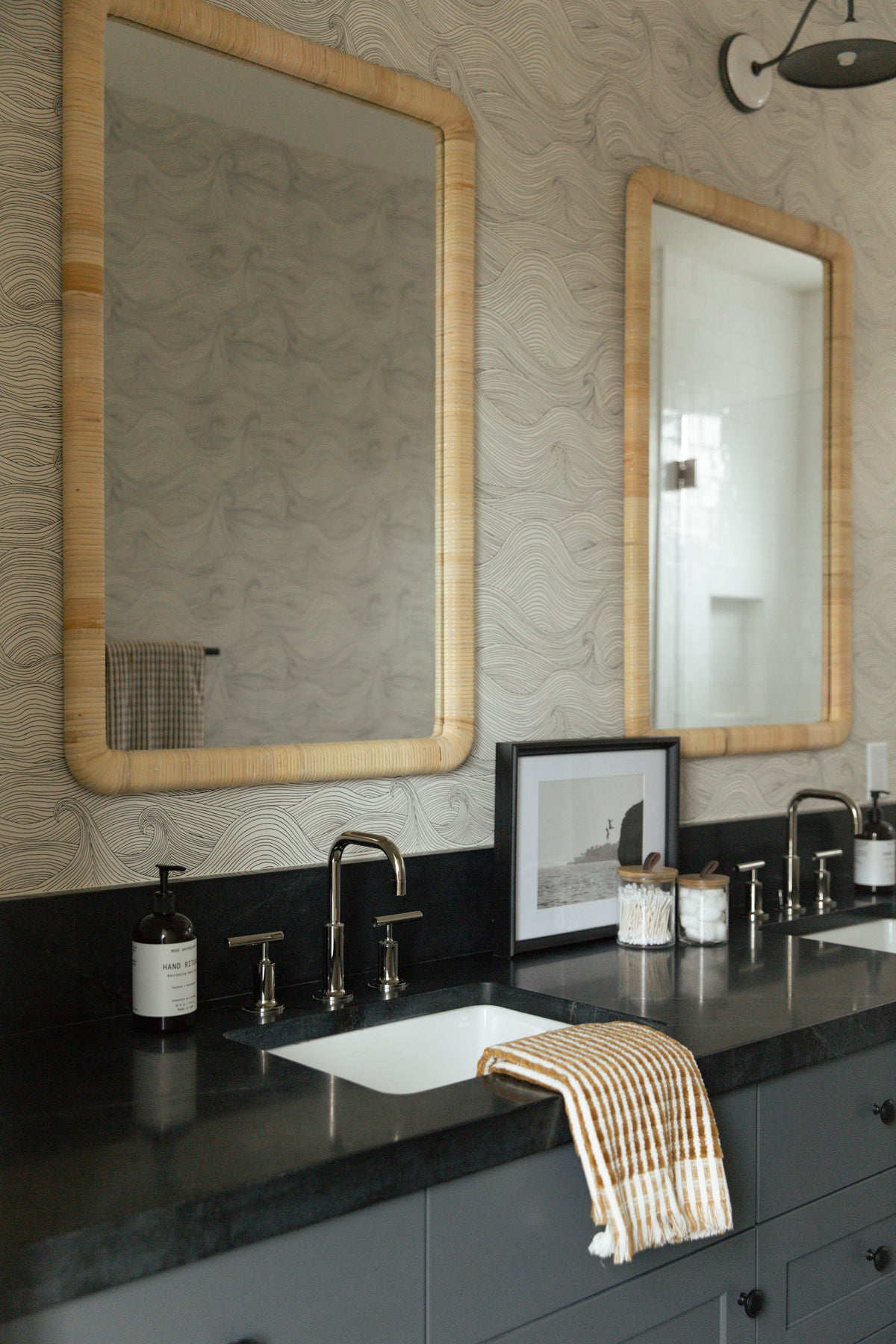 A beautifully styled coastal bathroom double vanity featuring dark grey cabinetry, solid dark countertops, dual wall-mounted faucets, and two arched rattan mirrors set against wave wallpaper.