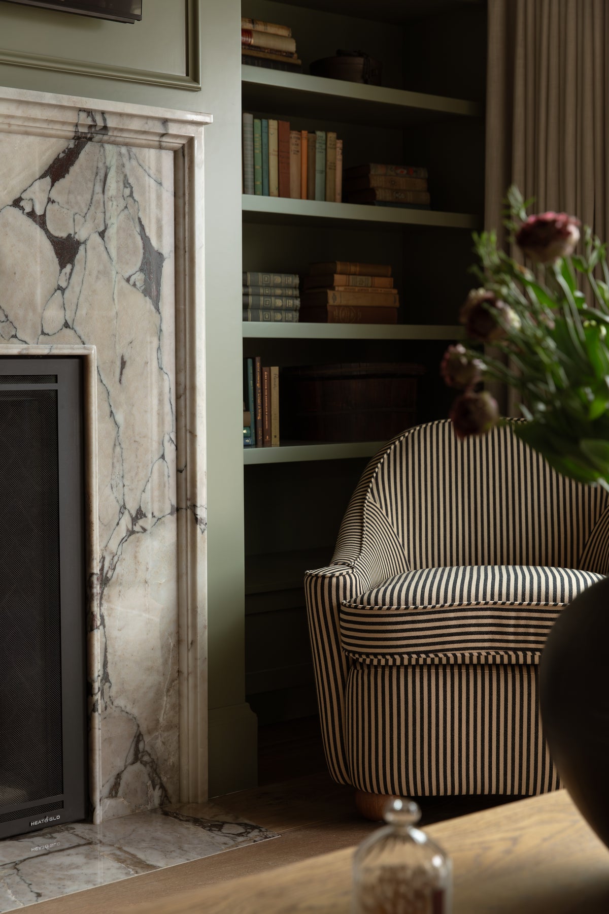 Cozy reading nook featuring a white veined marble fireplace, built-in sage green bookshelves, and a black-and-tan striped armchair.