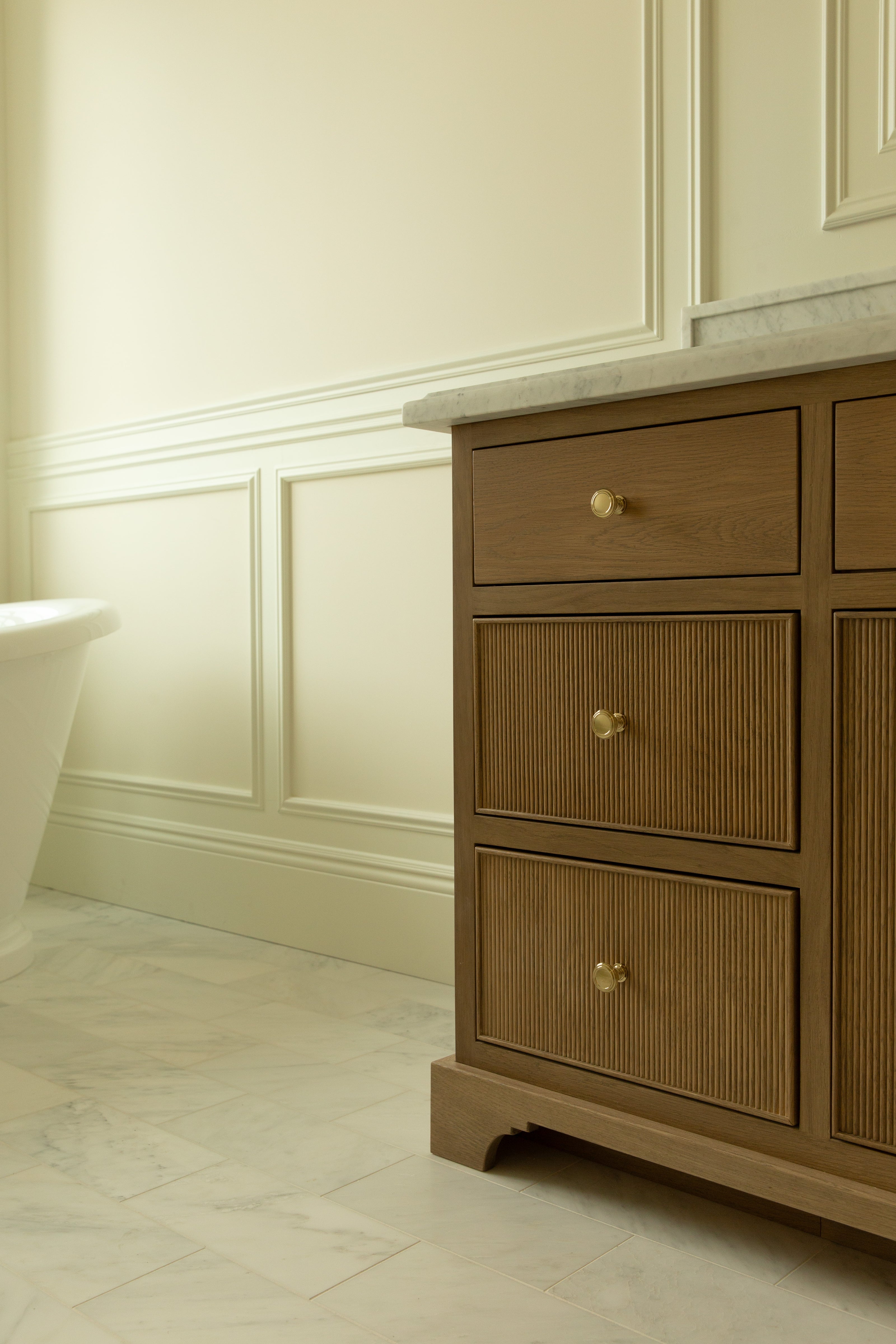 Close-up of ribbed wood bathroom cabinetry drawers with brass knobs, topped with a marble counter, with a glimpse of a white freestanding soaking tub in the background and wainscoting on the walls. 