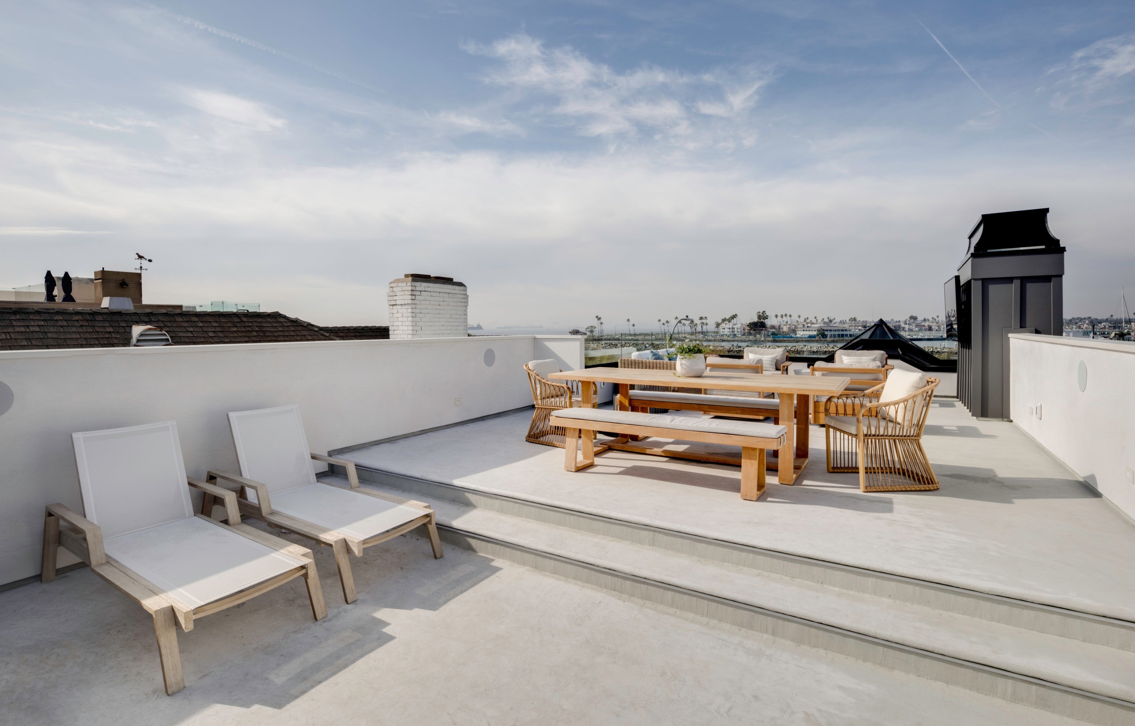 Luxury rooftop patio designed by Seal Beach interior design studio B Farm Design. This ocean-view deck features separate zones for sunbathing with white sling-back chaise lounges and outdoor dining, complete with a long teak table and matching bench seating set against the coastal landscape.
