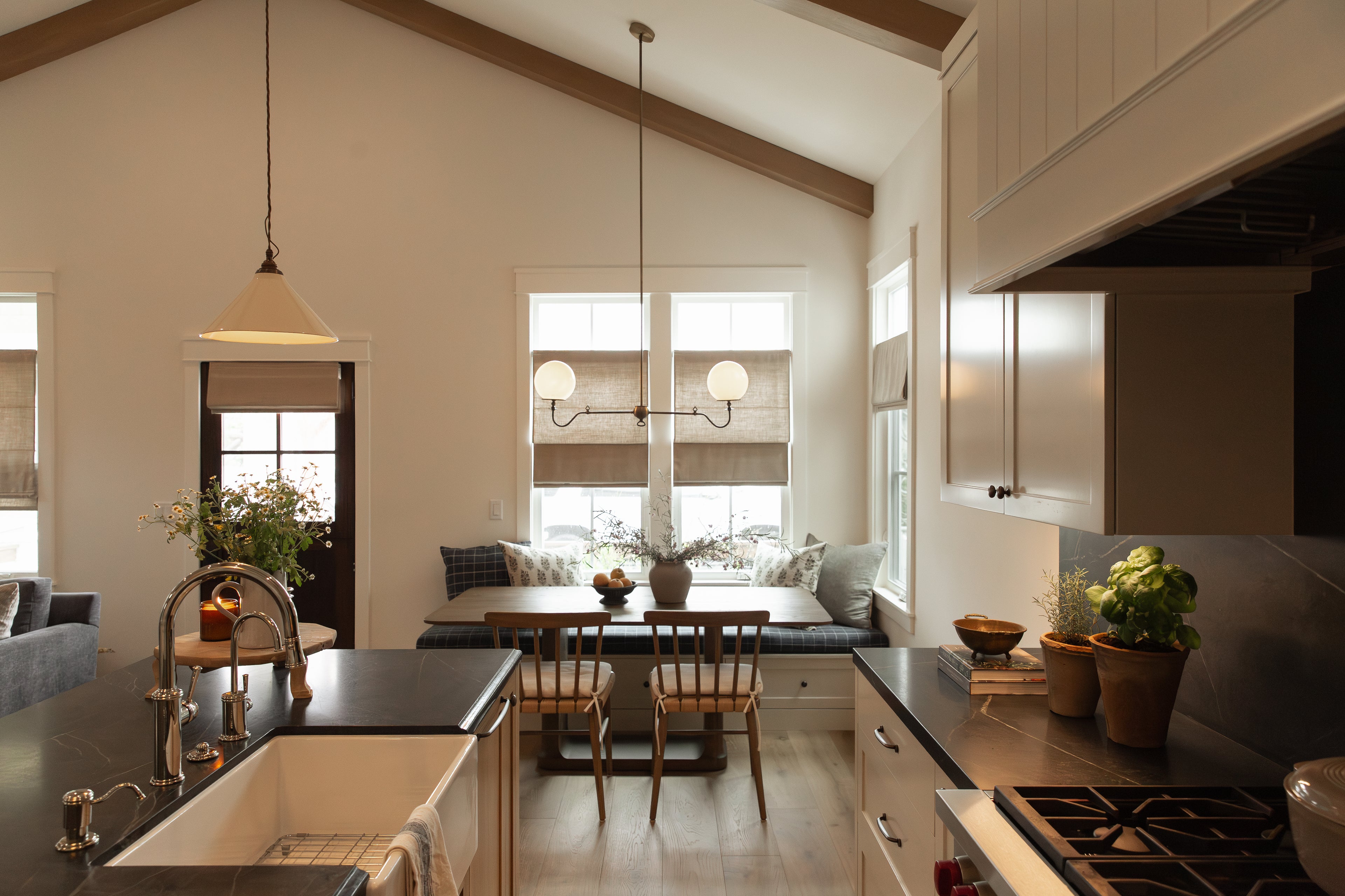 A wide view of an open-concept kitchen and dining space in a remodeled Seal Beach bungalow. The transitional coastal design features vaulted ceilings with exposed wood beams, a custom built-in window banquette, a kitchen island with dark slab countertops and a farmhouse sink, and elegant brass and glass pendant lighting.