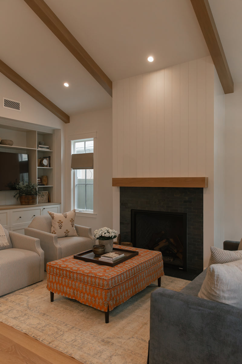 Wide view of a remodeled classic bungalow living room in Seal Beach, CA, featuring a custom white shiplap fireplace with a black tile surround, exposed wood ceiling beams, tailored furnishings, and custom built-in cabinetry.