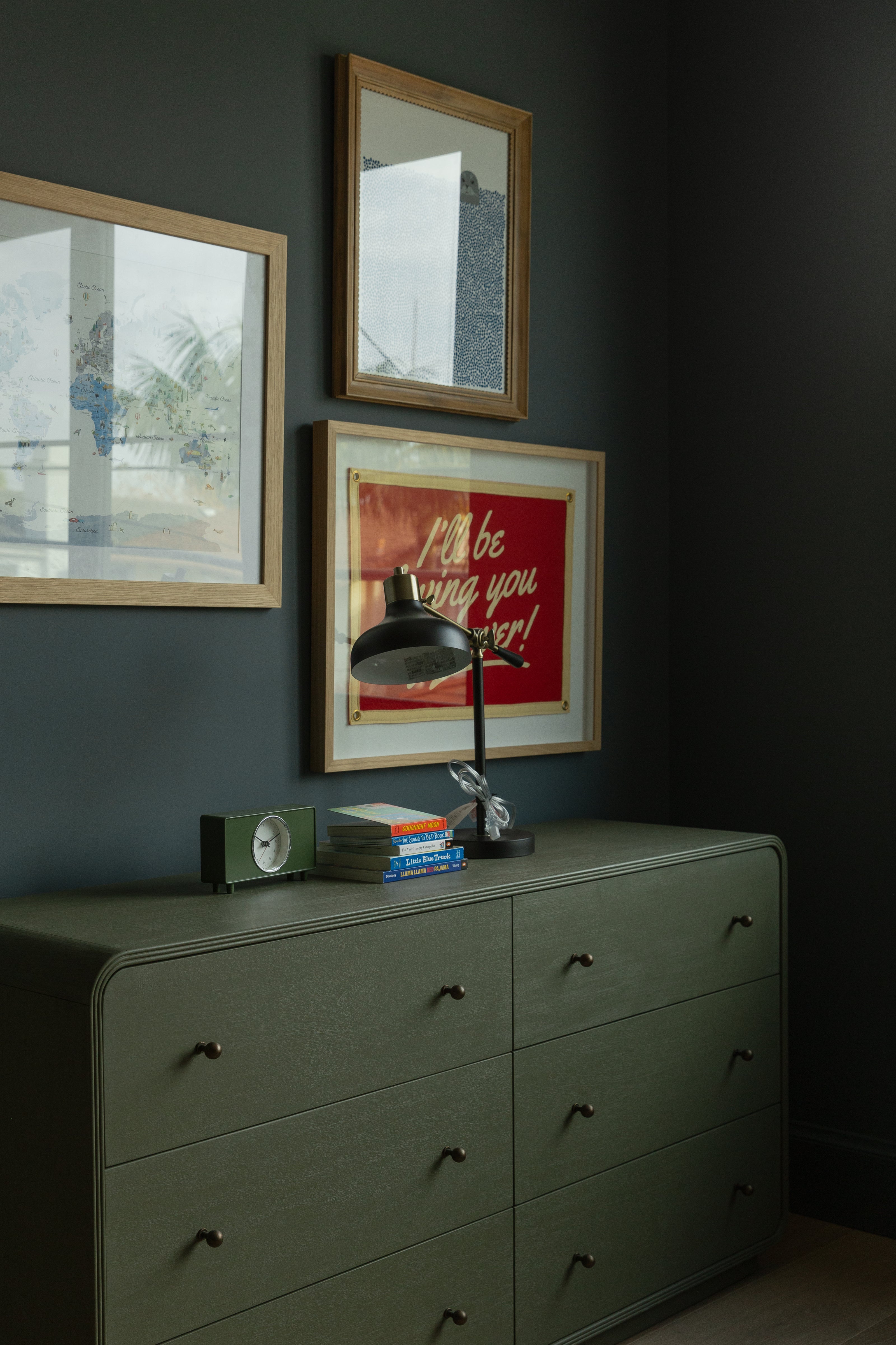 A beautifully styled moody kids room in a Seal Beach custom home, showcasing an olive green rounded dresser, dark paneled walls, a black desk lamp, and a curated gallery wall.