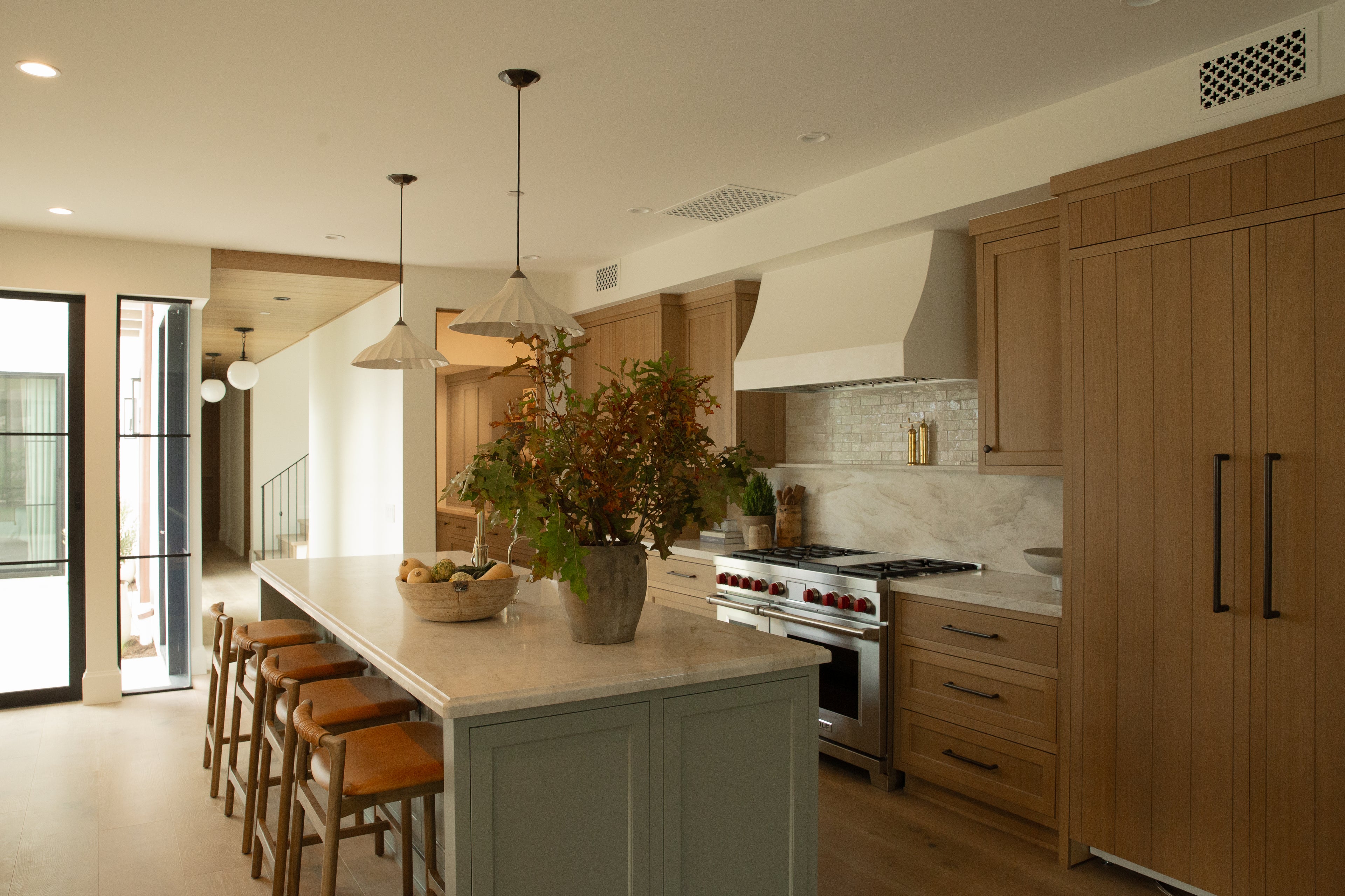 A wide view of an open-concept kitchen in a Seal Beach custom home, featuring a large seating island, warm oak cabinetry, custom lighting, and a view down a bright hallway with classic arched doorways.