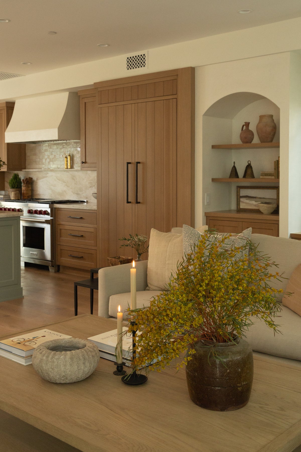 An open-concept transitional living space showcasing the seamless design flow from a cozy living room with an arched display niche into a warm, light oak custom kitchen.