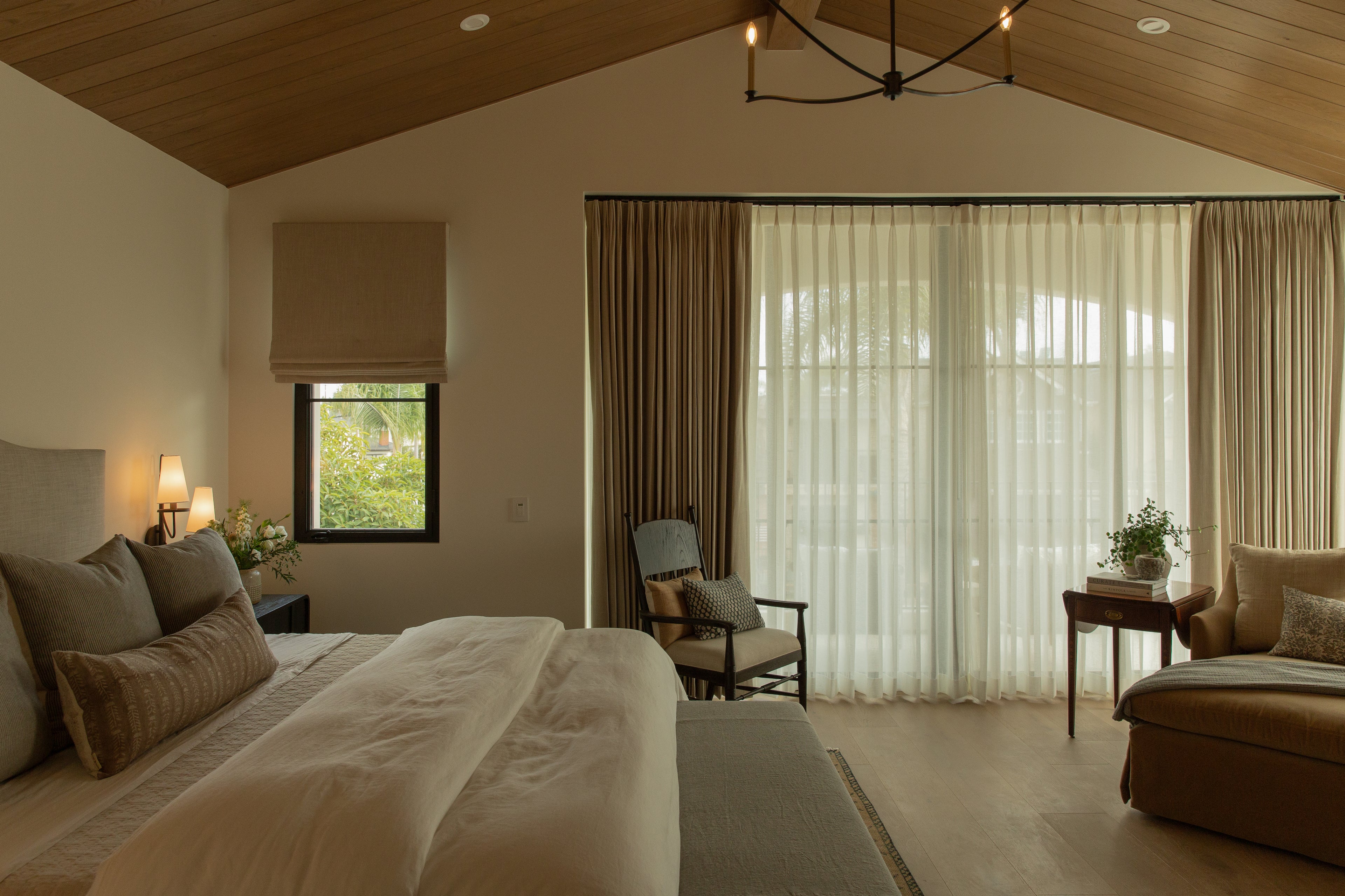A luxurious primary bedroom in a ground-up Old Town Seal Beach custom home, showcasing a vaulted wood plank ceiling, a dark iron chandelier, custom layered drapery, and a beautifully upholstered bed.
