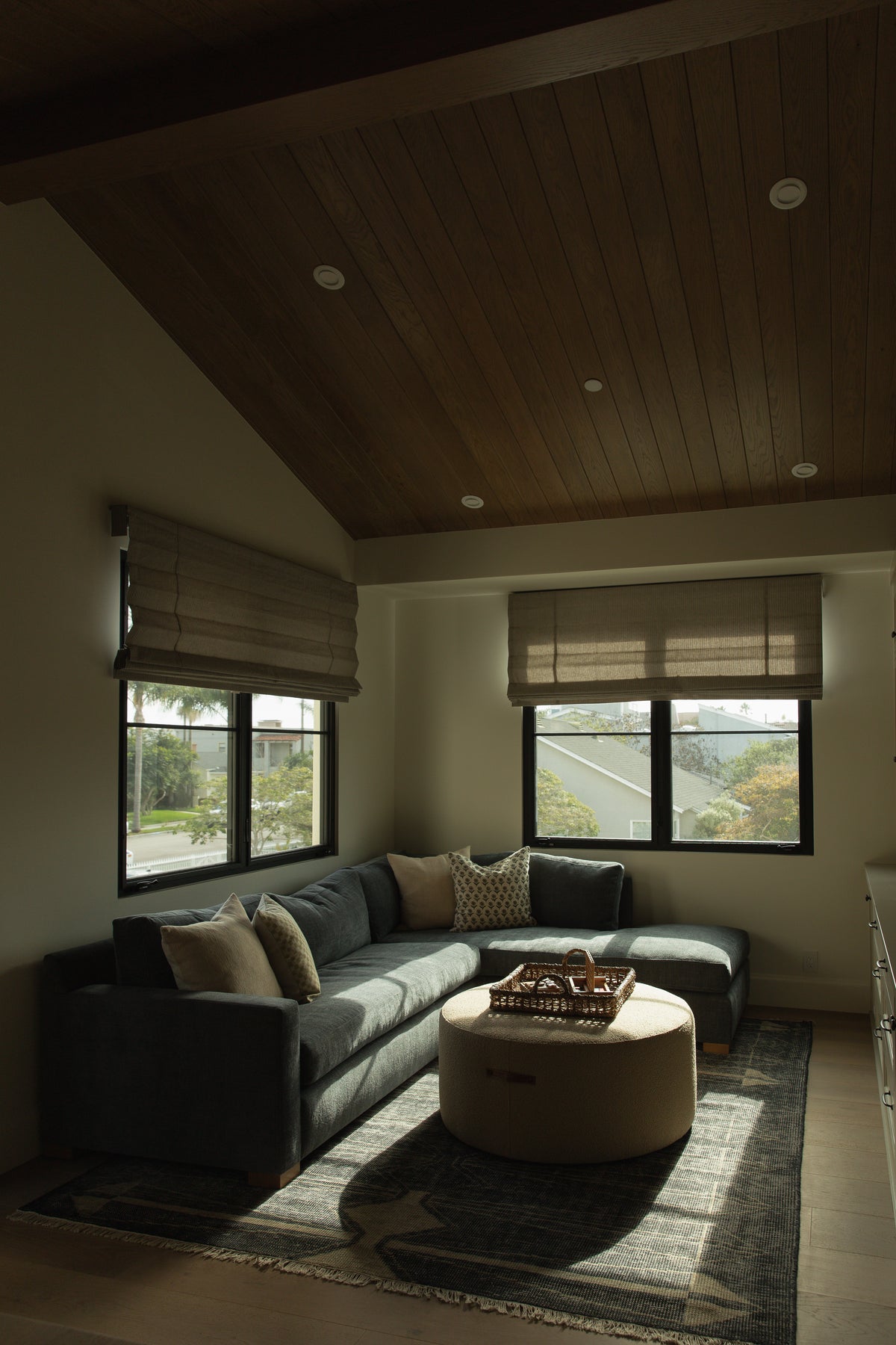 A sunlit bonus room in a Seal Beach custom home, showcasing a striking vaulted wood plank ceiling, custom patterned roman shades on black-framed windows, and a comfortable corner seating area.