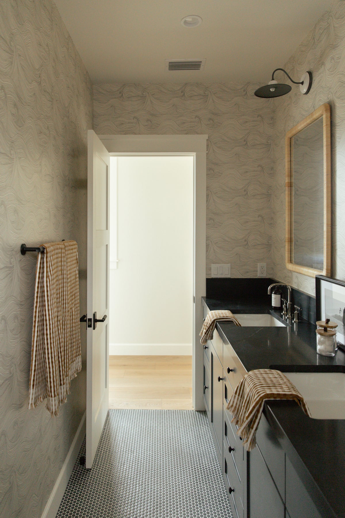 Looking through an open white door into a coastal farmhouse bathroom in Surfside, CA, featuring a dark double vanity, grey penny tile flooring, and subtle wave patterned wallpaper.