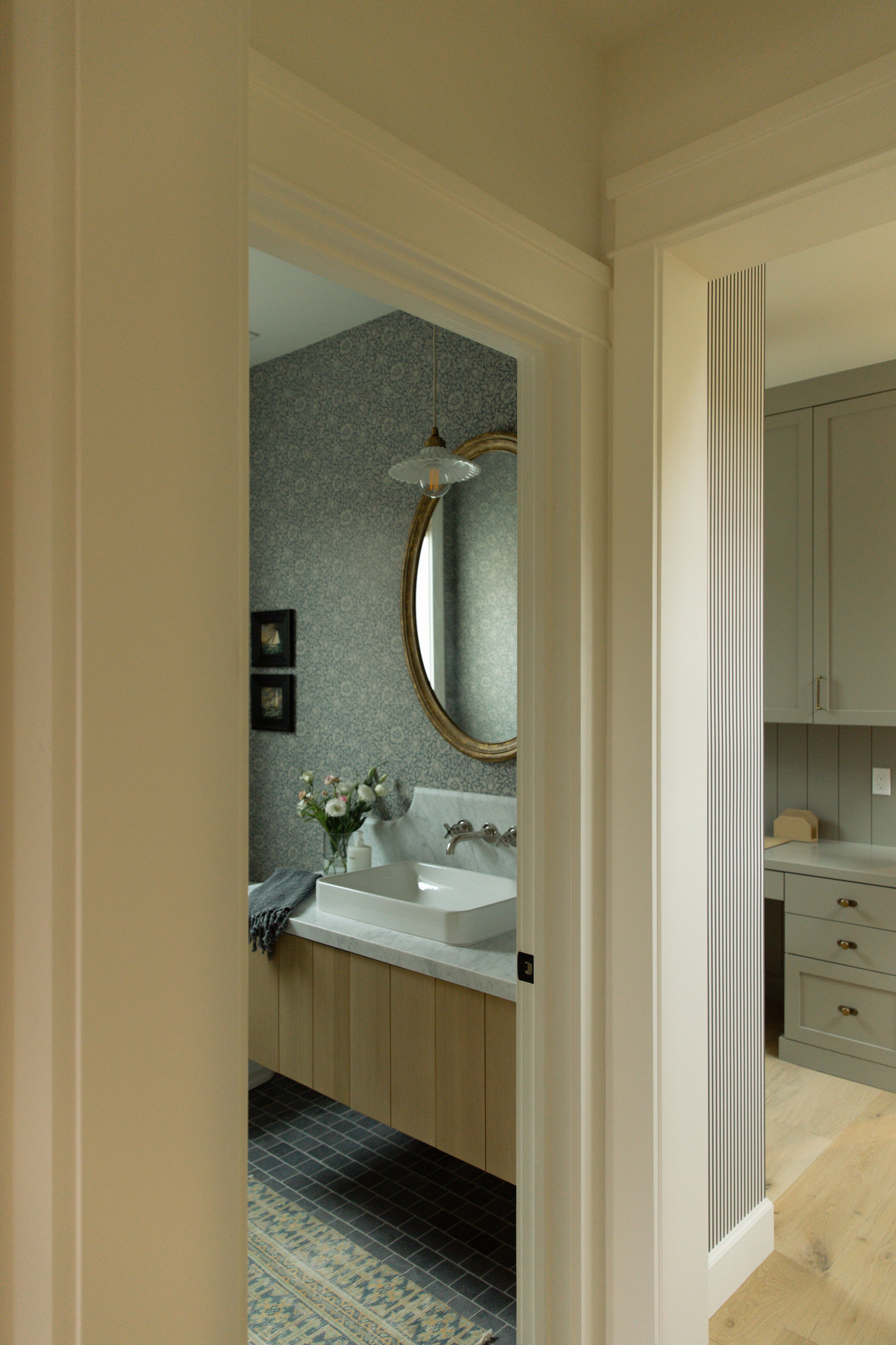 A peek from the hallway into a Surfside coastal farmhouse powder room featuring a light wood vanity, beautiful blue floral wallpaper, and a marble countertop.