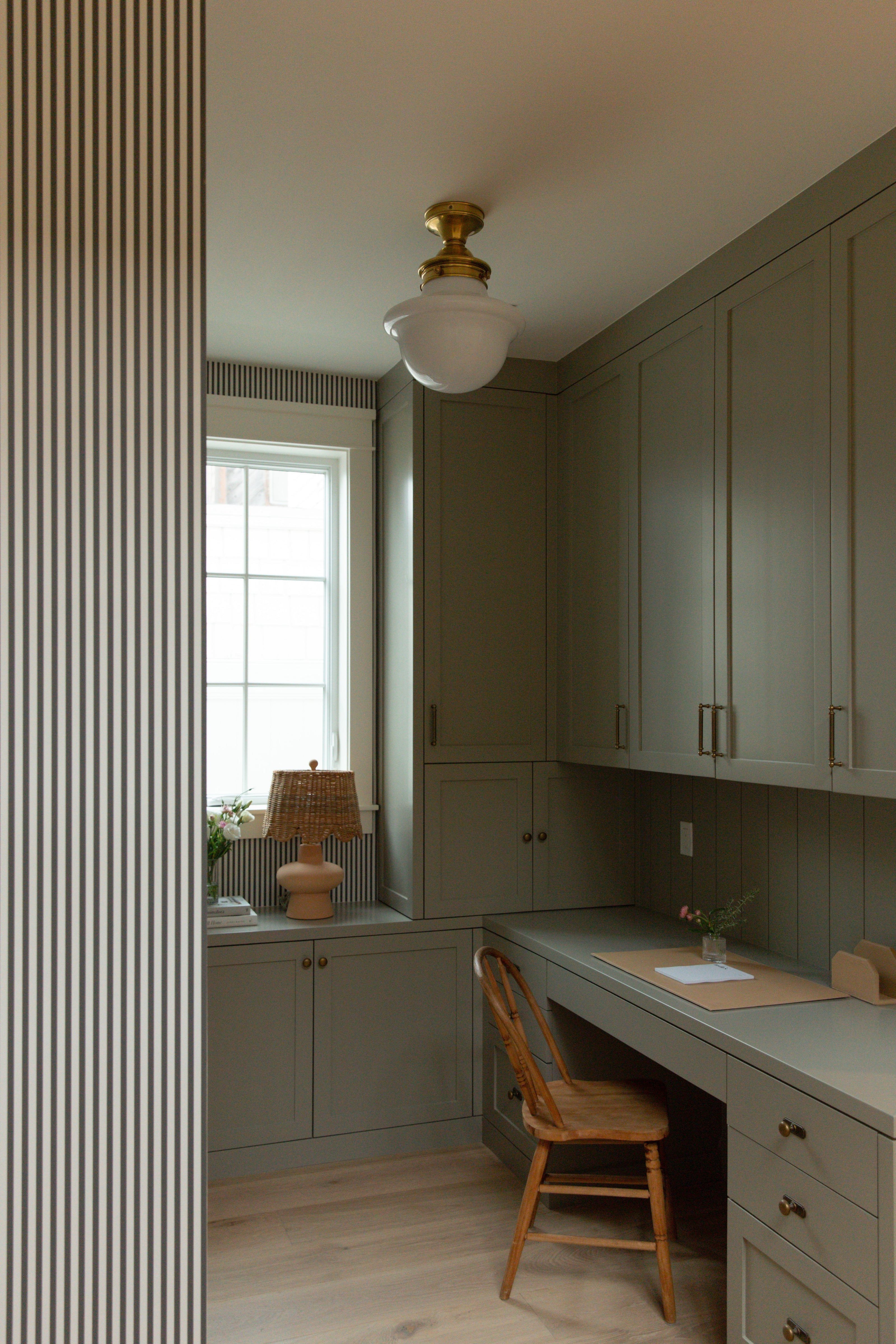A coastal farmhouse home office in a Surfside custom build, featuring floor-to-ceiling grey-green built-in cabinetry, classic striped wallpaper, and a vintage wooden desk chair.