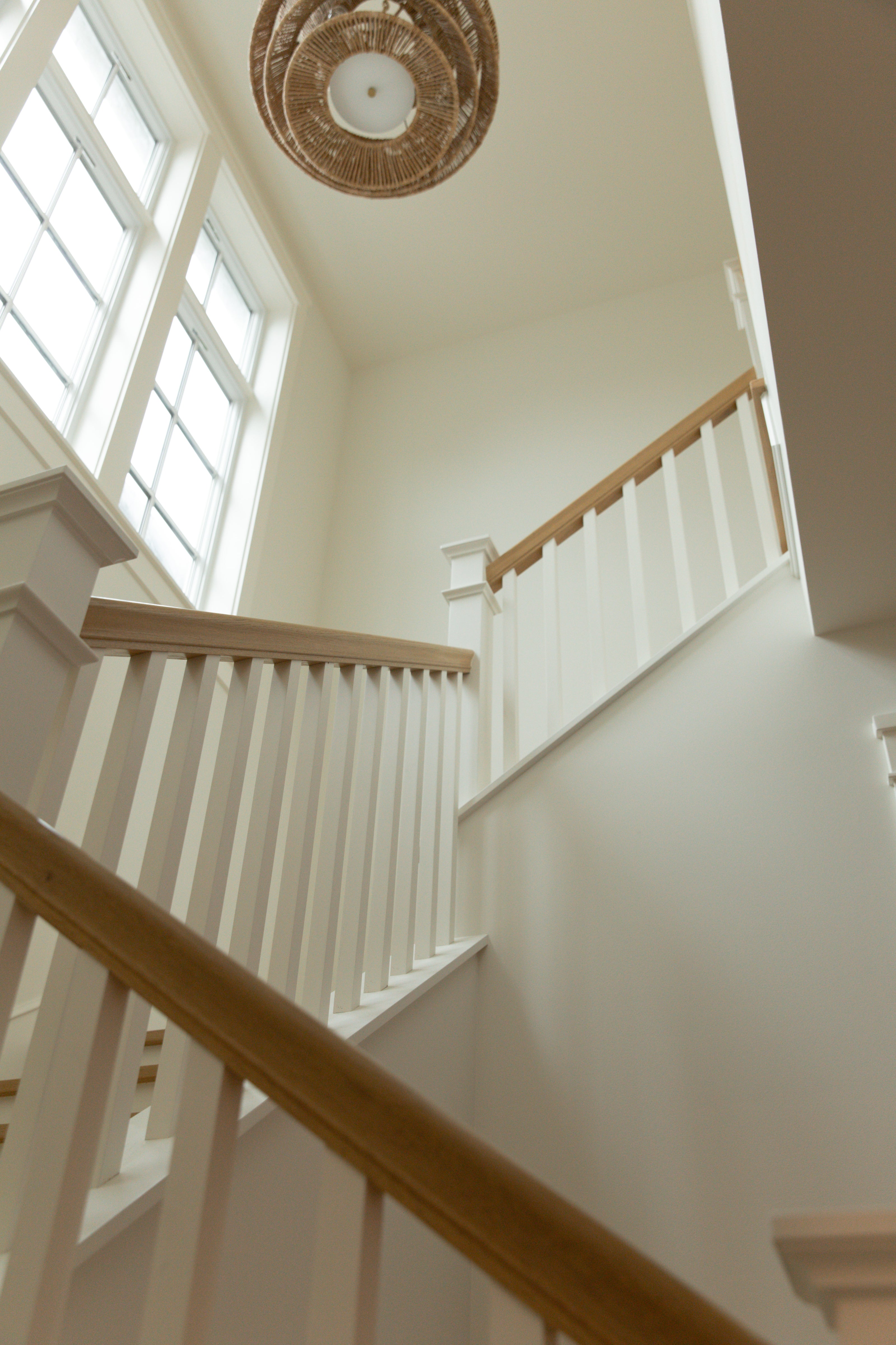 Looking up a grand staircase in a Surfside custom home, featuring classic white balusters, a light oak handrail, and a beautiful woven rattan tiered chandelier.