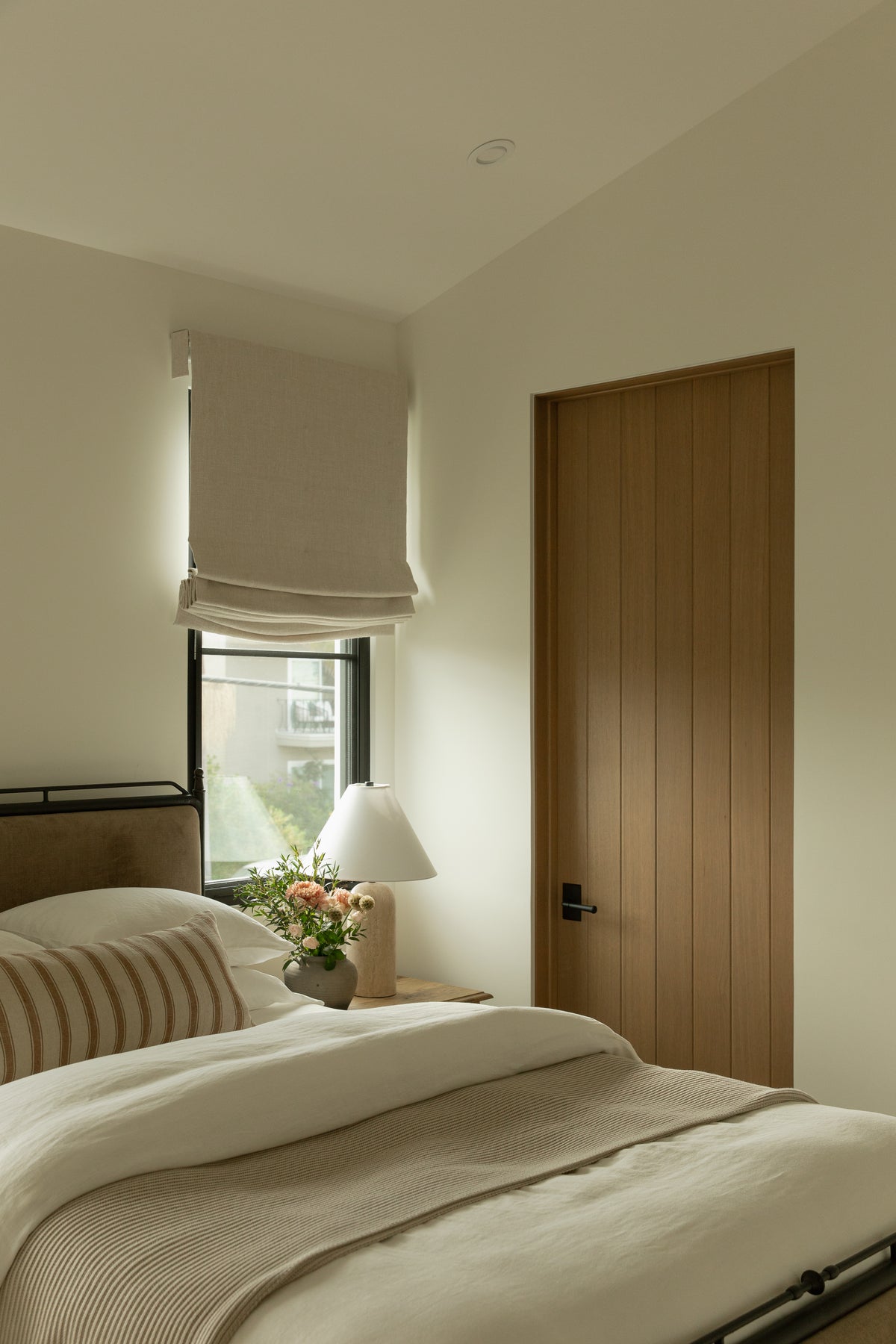 A beautifully appointed transitional bedroom highlighting a custom vertical-plank wood door, an iron bed frame with neutral linens, and a tailored roman shade over a black-framed window.
