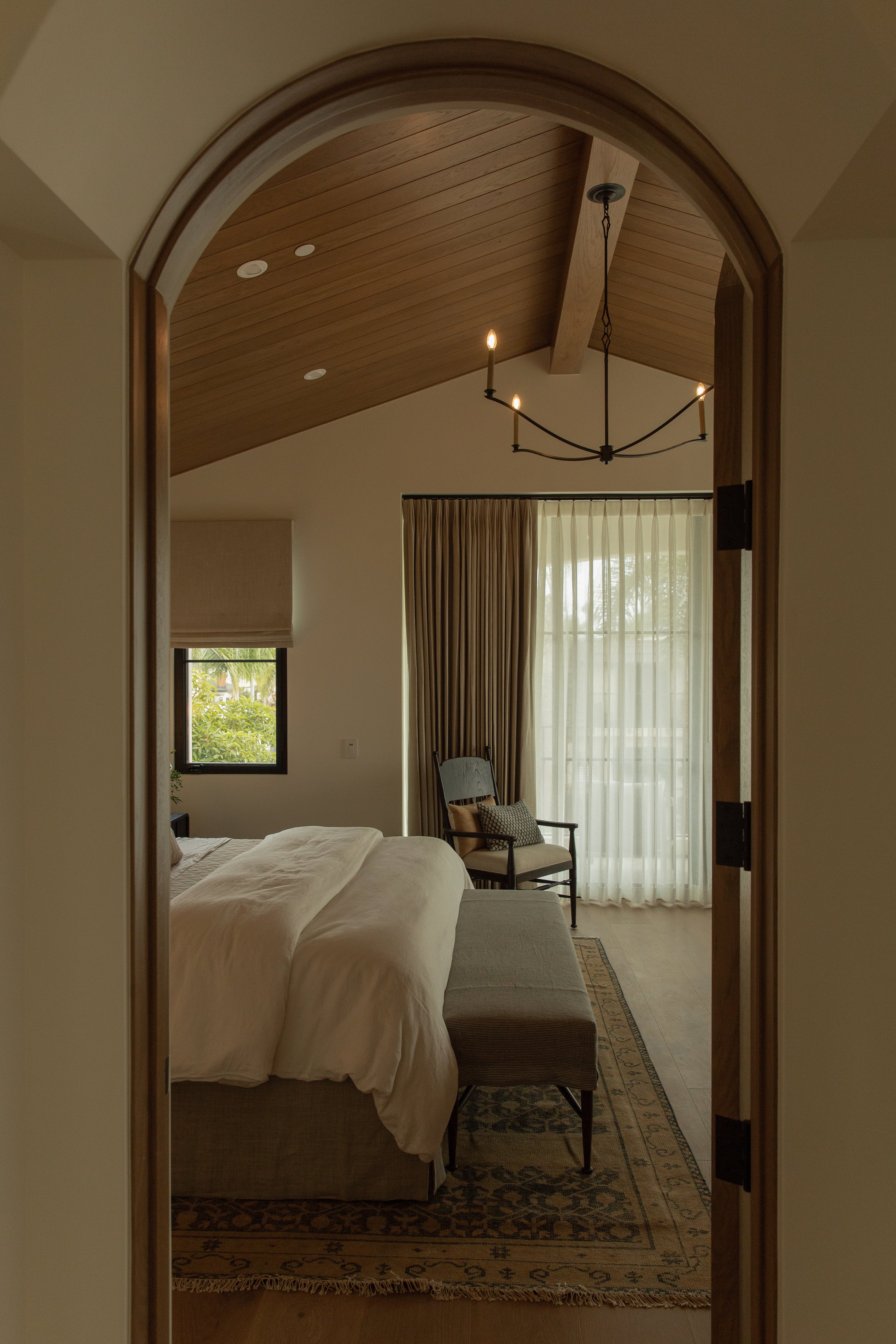 A view through a classic arched doorway into a transitional primary bedroom featuring a warm wood-paneled vaulted ceiling, expansive glass doors with sheer drapery, and a vintage accent chair.