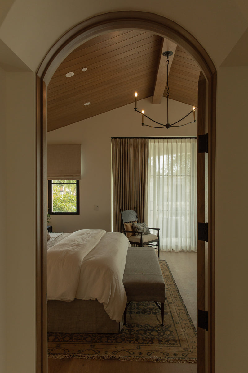 A view through a classic arched doorway into a transitional primary bedroom featuring a warm wood-paneled vaulted ceiling, expansive glass doors with sheer drapery, and a vintage accent chair.