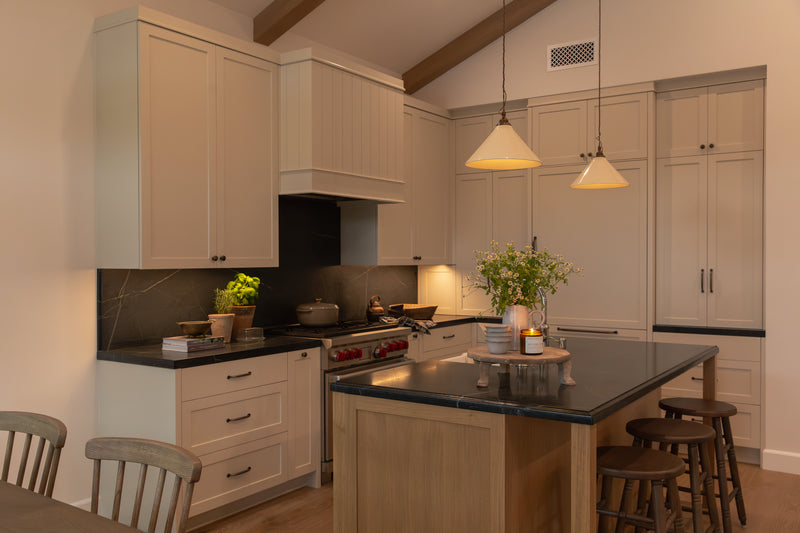 A fully redesigned classic bungalow kitchen in Orange County highlighting custom white shaker cabinetry, a matching dark solid surface countertop and backsplash, classic cone pendant lights, and a custom range hood.