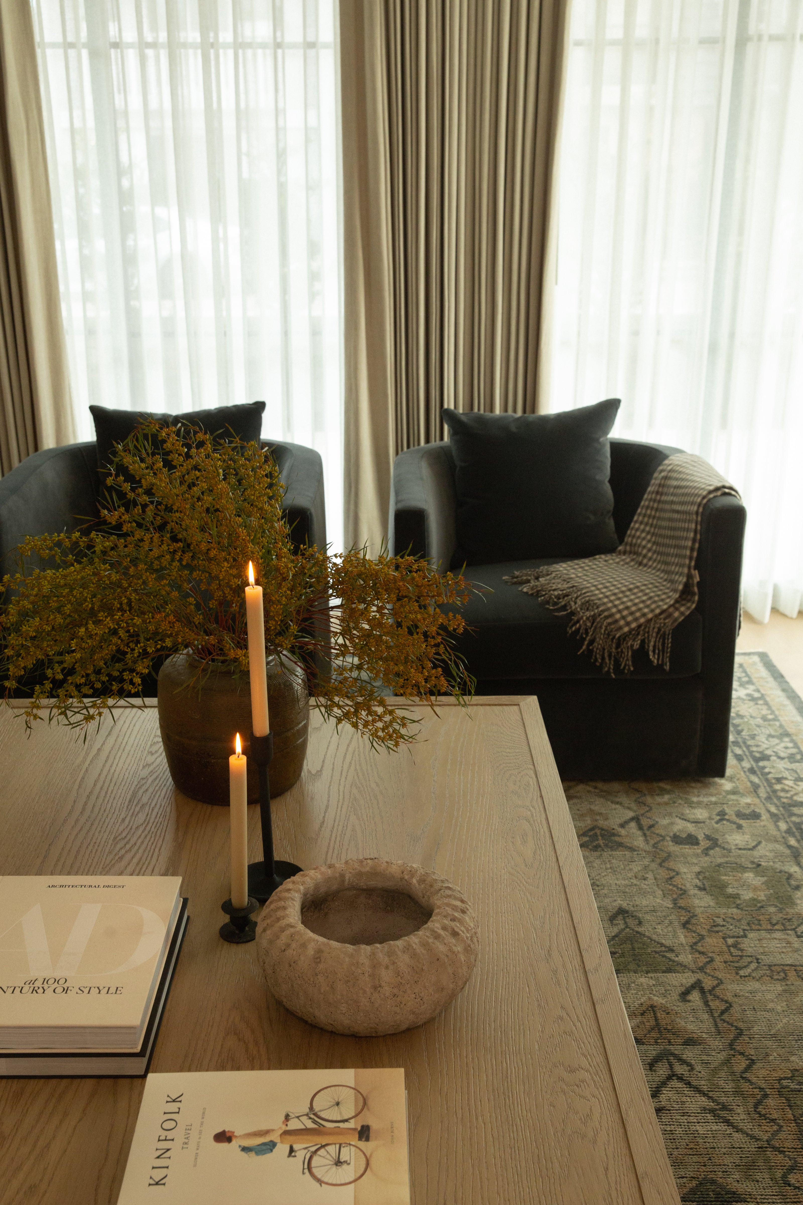 Close-up of coffee table styling in a transitional living room, featuring architectural design books, taper candles in iron holders, and a rustic stone bowl, with two dark velvet swivel chairs in the background.