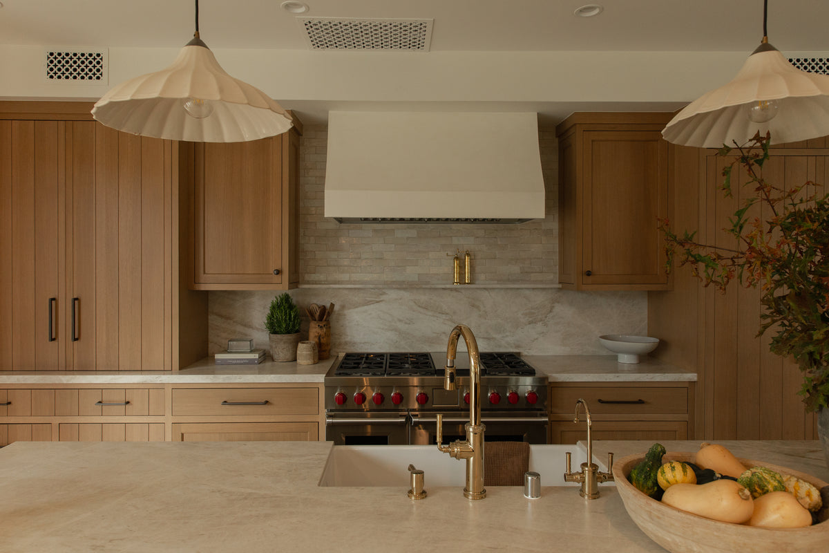 A beautifully styled transitional kitchen island featuring a polished brass bridge faucet, a white farmhouse sink, fluted ceramic pendant lights, and light oak shaker cabinetry.