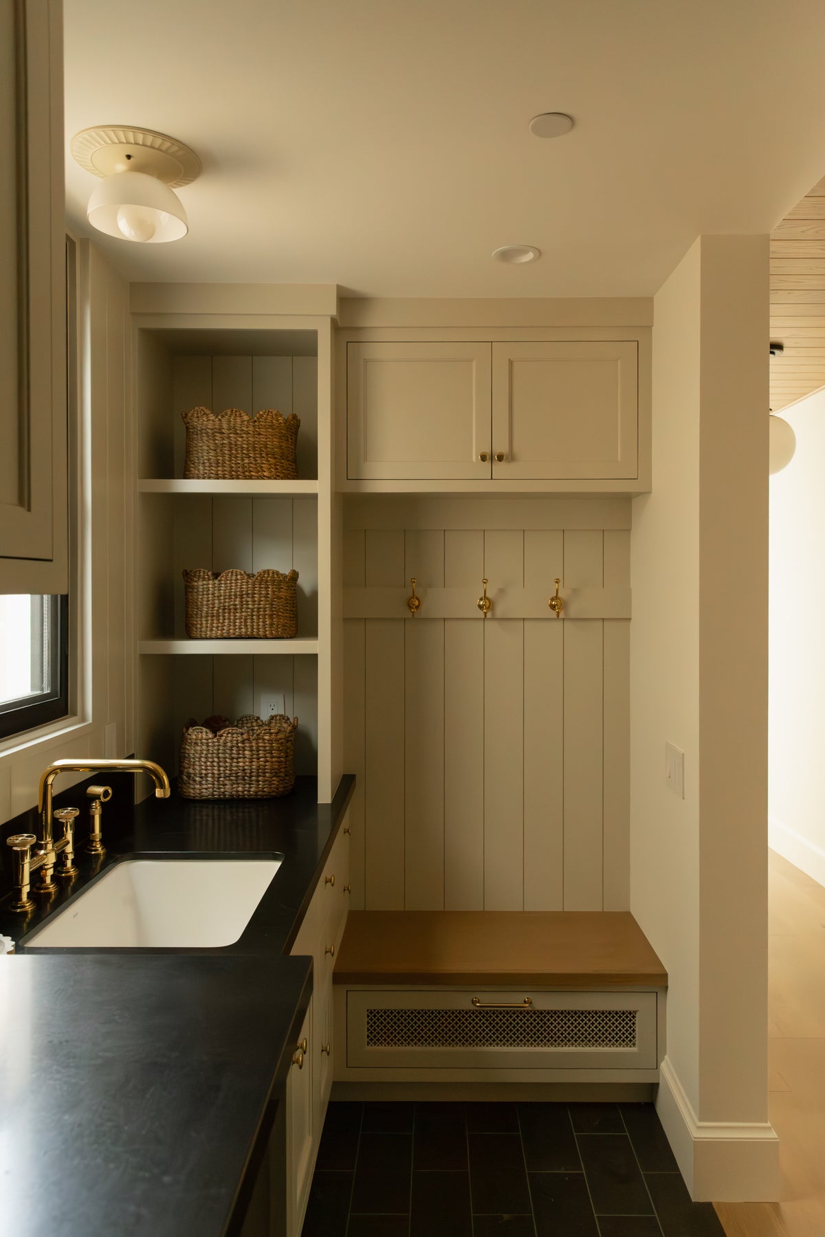 A functional and beautiful transitional laundry and mudroom featuring custom off-white cabinetry, a dark soapstone utility sink, unlacquered brass bridge faucet, and a bench with black and white checked upholstery.