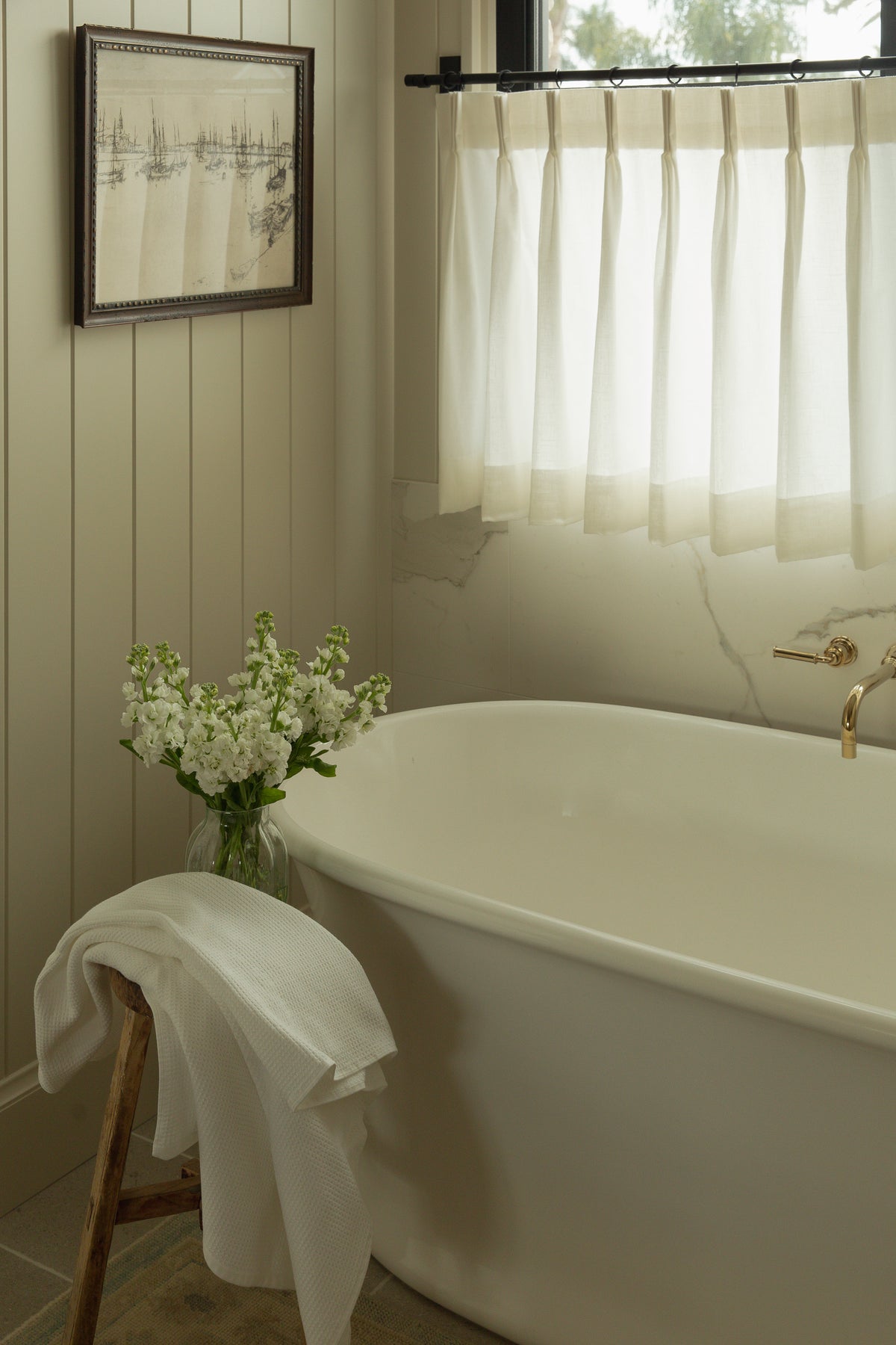 A serene transitional bathroom corner showcasing a white freestanding tub, a wall-mounted brass tub filler, vertical shiplap paneling, and a black framed window dressed with light linen cafe curtains.