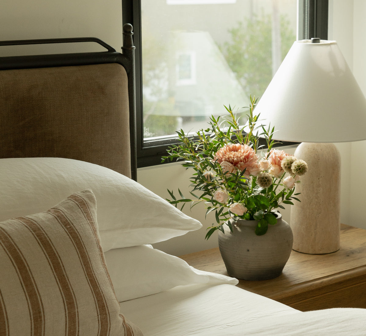 Close-up of a warm wood nightstand in a transitional bedroom, beautifully styled with a solid travertine table lamp, fresh florals in a rustic ceramic vase, and layered neutral bedding.