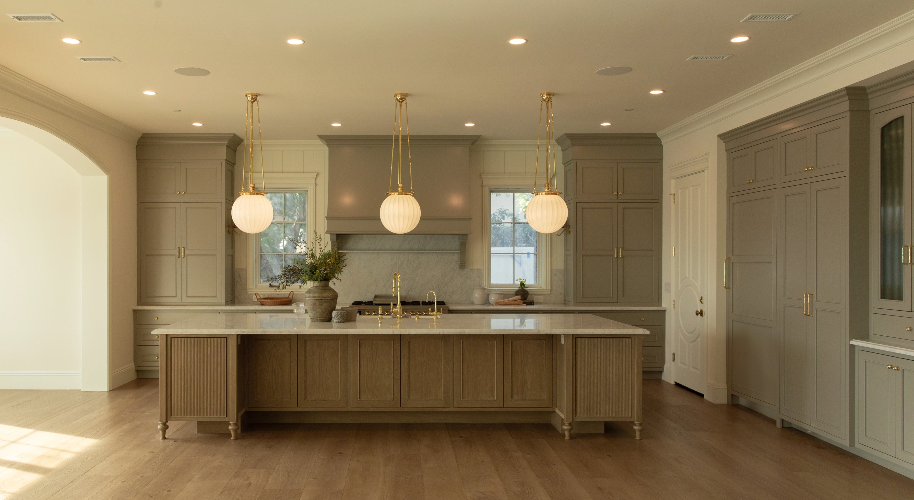View through an arched doorway into a custom Tudor kitchen remodel in Rossmoor, featuring a large fluted white oak island, brass globe pendant lights, and wide plank wood flooring.