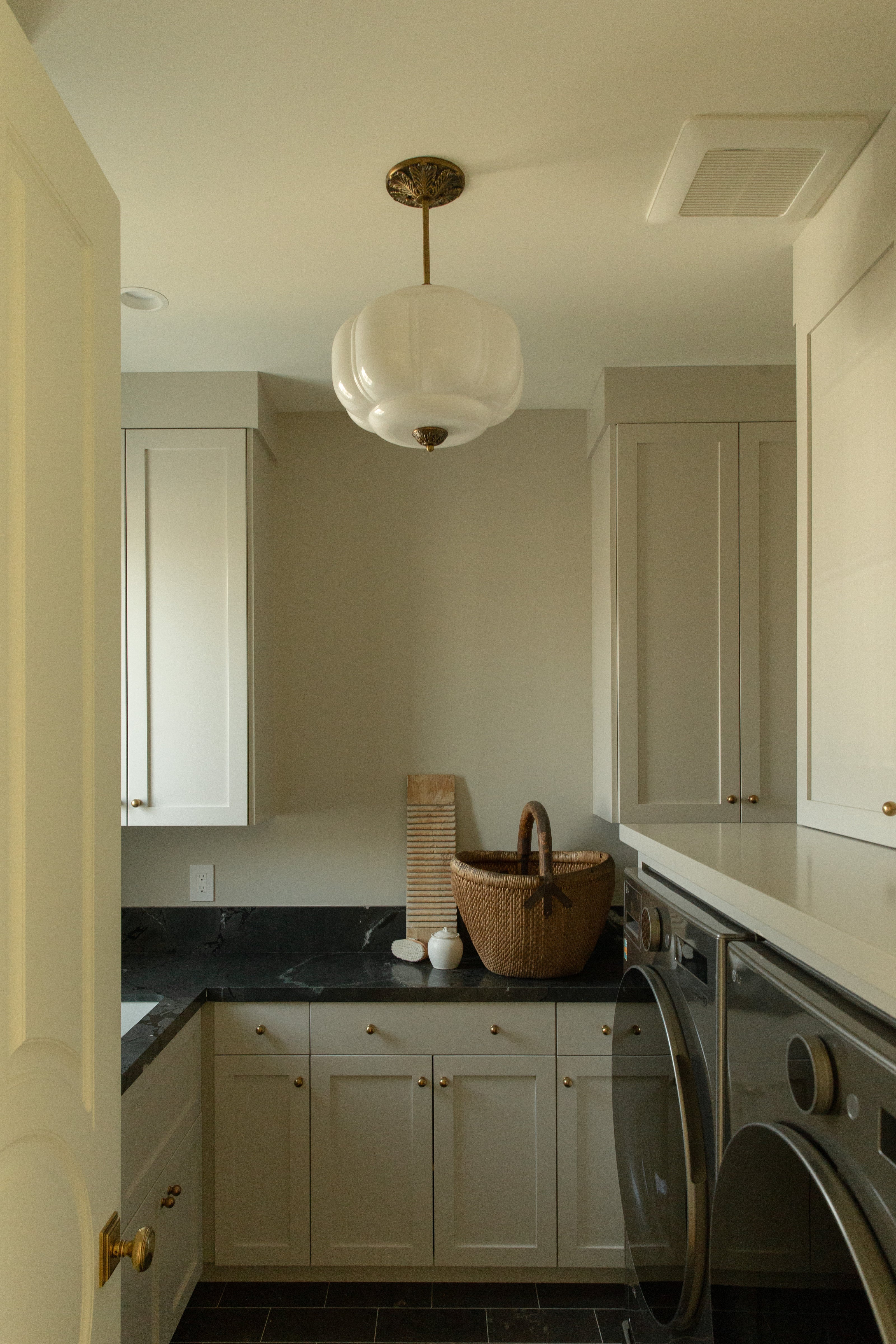 Custom Tudor-style laundry room featuring light cabinetry with brass knobs, dark stone countertops, a vintage-style scalloped pendant light, and front-loading appliances.