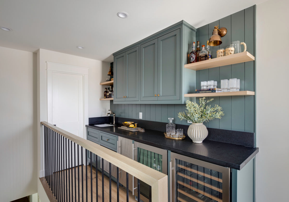 A custom upstairs wet bar by B Farm Design. Features sage green cabinetry, a vertical shiplap backsplash, and light wood floating shelves. The space includes dark countertops with a bar sink, brass wall sconces, and integrated wine and beverage refrigerators on an upstairs landing.